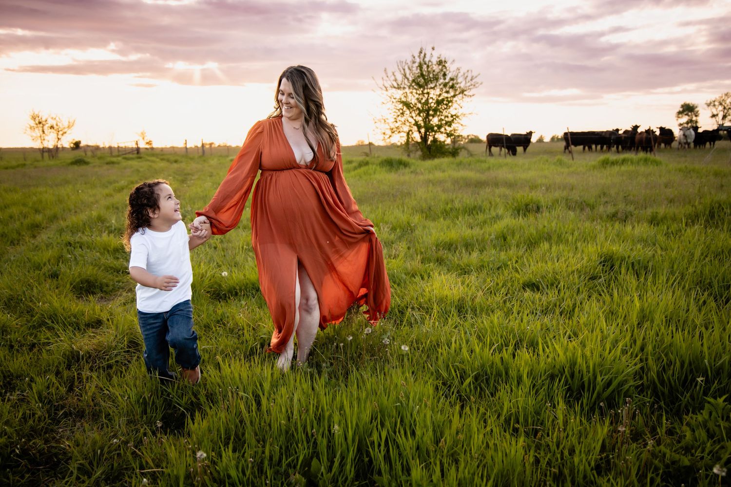Pregnant woman in orange dress walks with young child in green field at sunset with cattle grazing nearby.