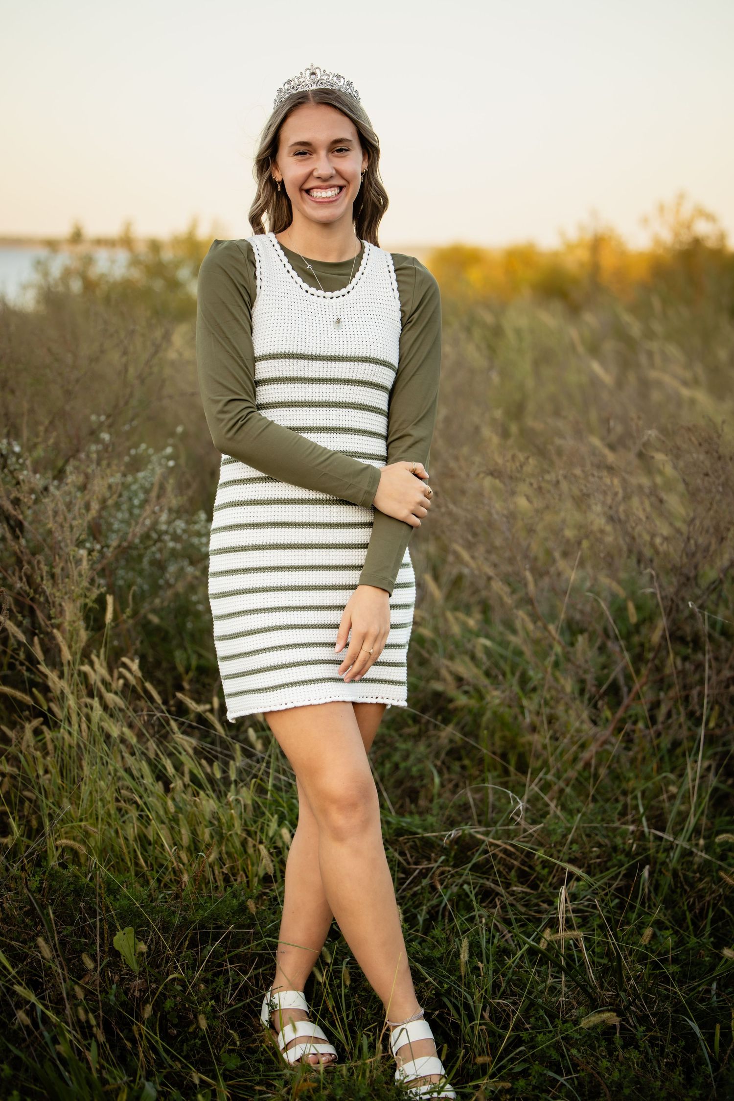 Smiling young woman in striped dress and tiara posing outdoors in golden field at sunset.
