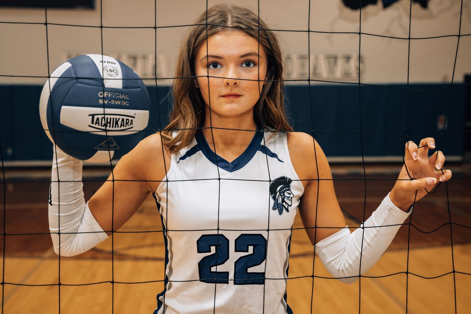 Female volleyball player wearing jersey number 22 holds a volleyball beside a net in a gymnasium.