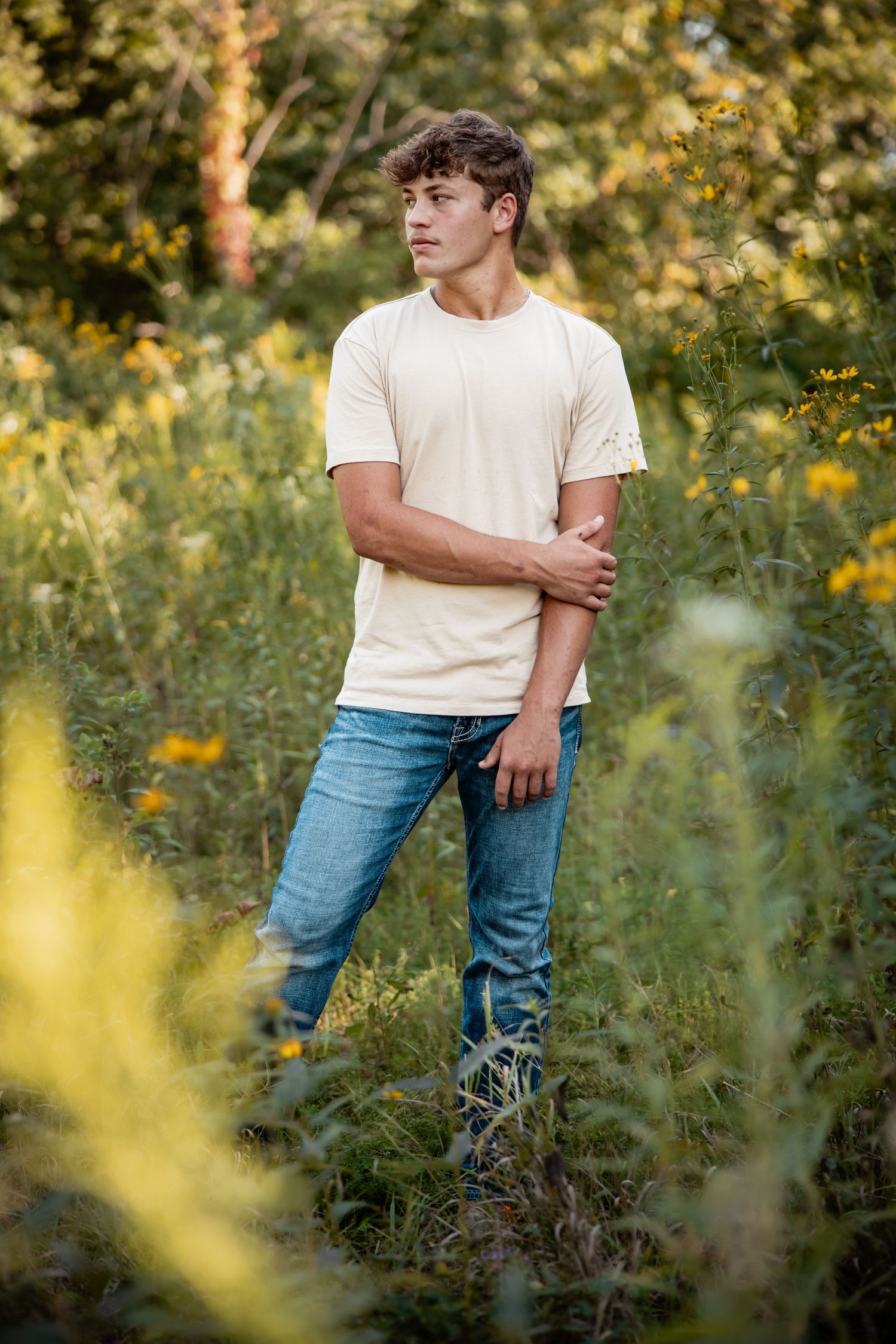 Young man in cream t-shirt and jeans standing among wildflowers and lush green foliage outdoors.