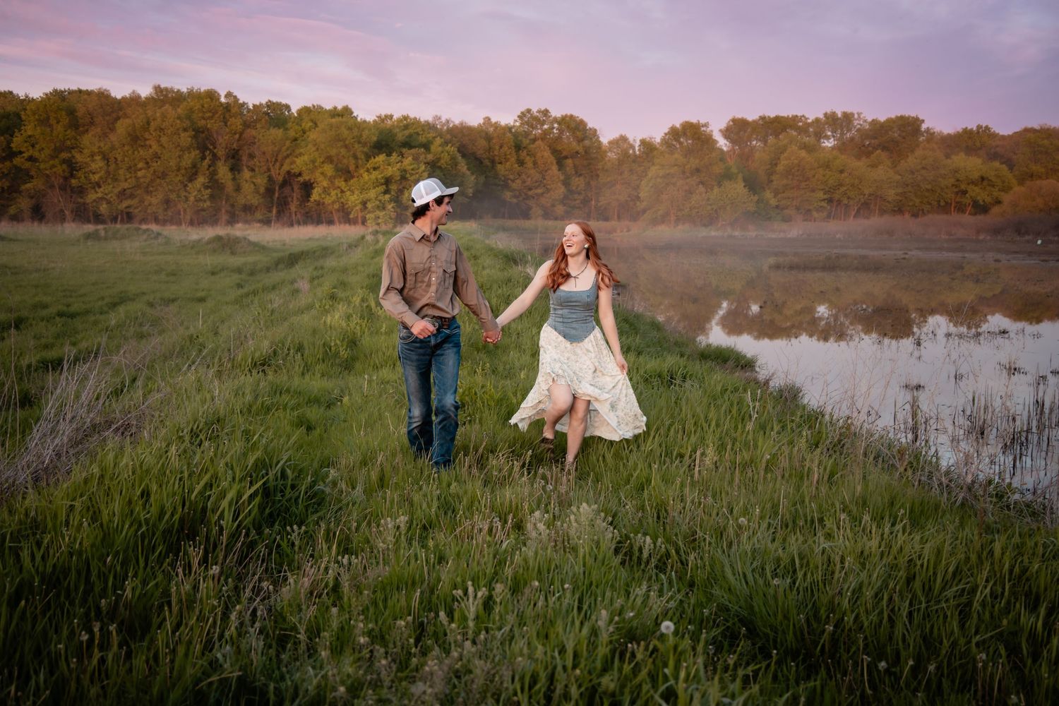 Couple walking hand in hand through tall grass near a pond at golden hour.