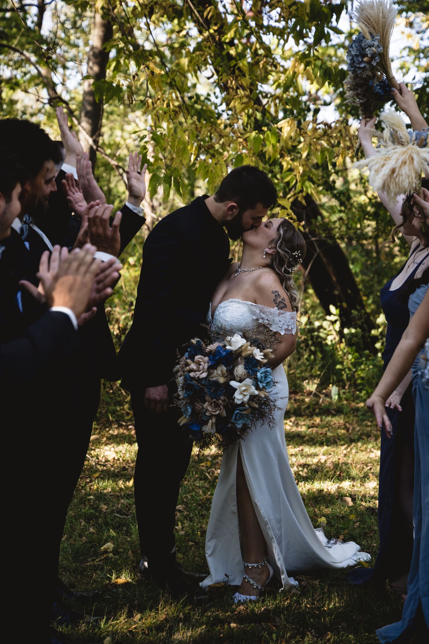 Newlyweds share a first kiss as wedding guests celebrate with pampas grass bouquets outdoors.