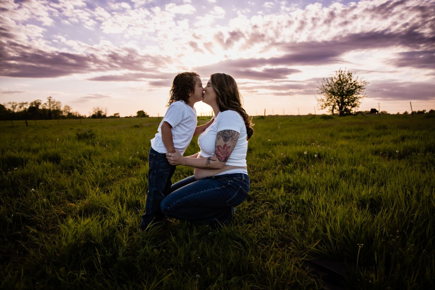 Two women share a kiss in a green field at sunset under a dramatic purple cloudy sky.