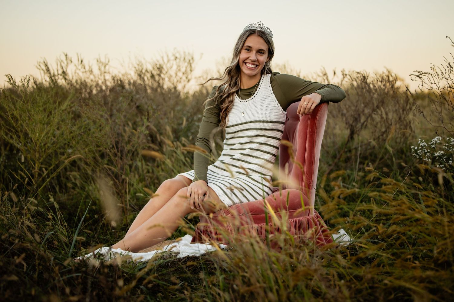 Smiling young woman in striped dress sitting on vintage chair surrounded by tall grass at golden hour.