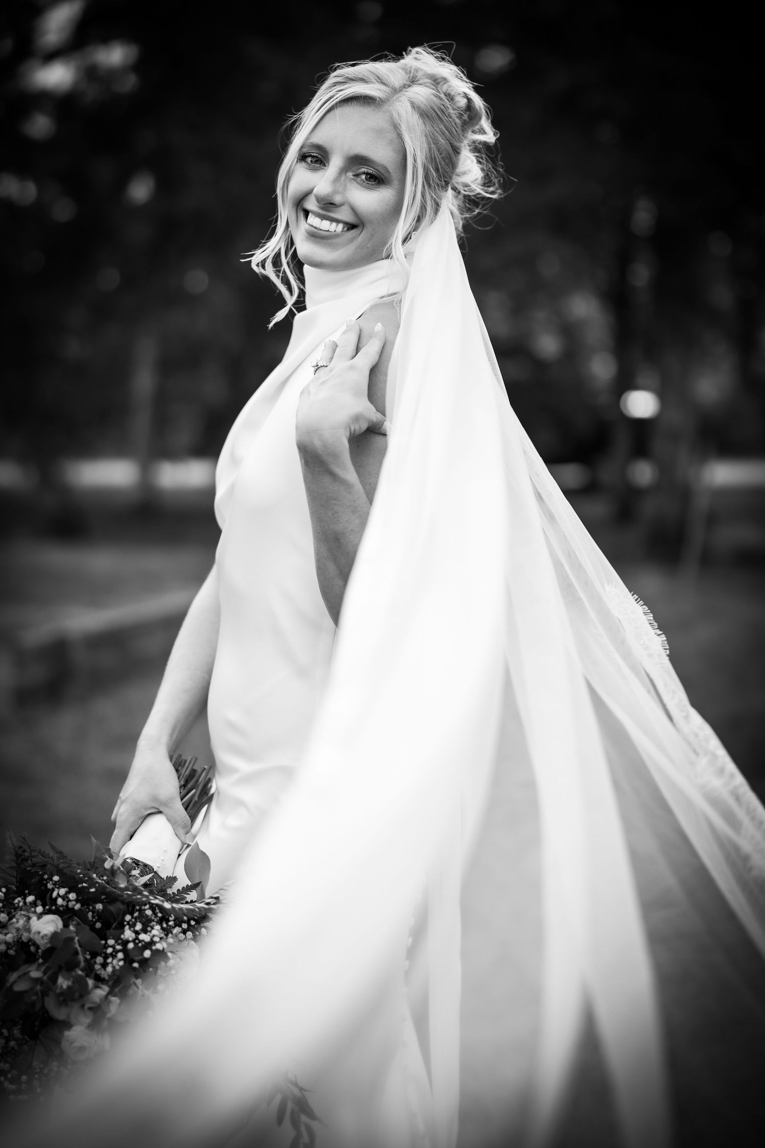 Black and white photo of a smiling bride in a white gown and flowing veil holding a bouquet outdoors.