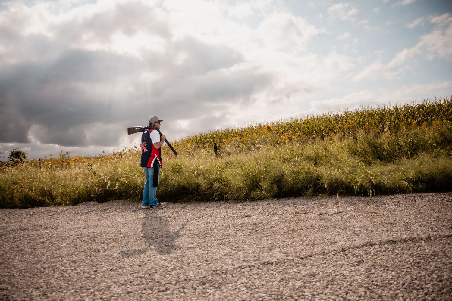 Person standing on rural road beside cornfield under dramatic cloudy sky, wearing backpack and spreading arms wide.