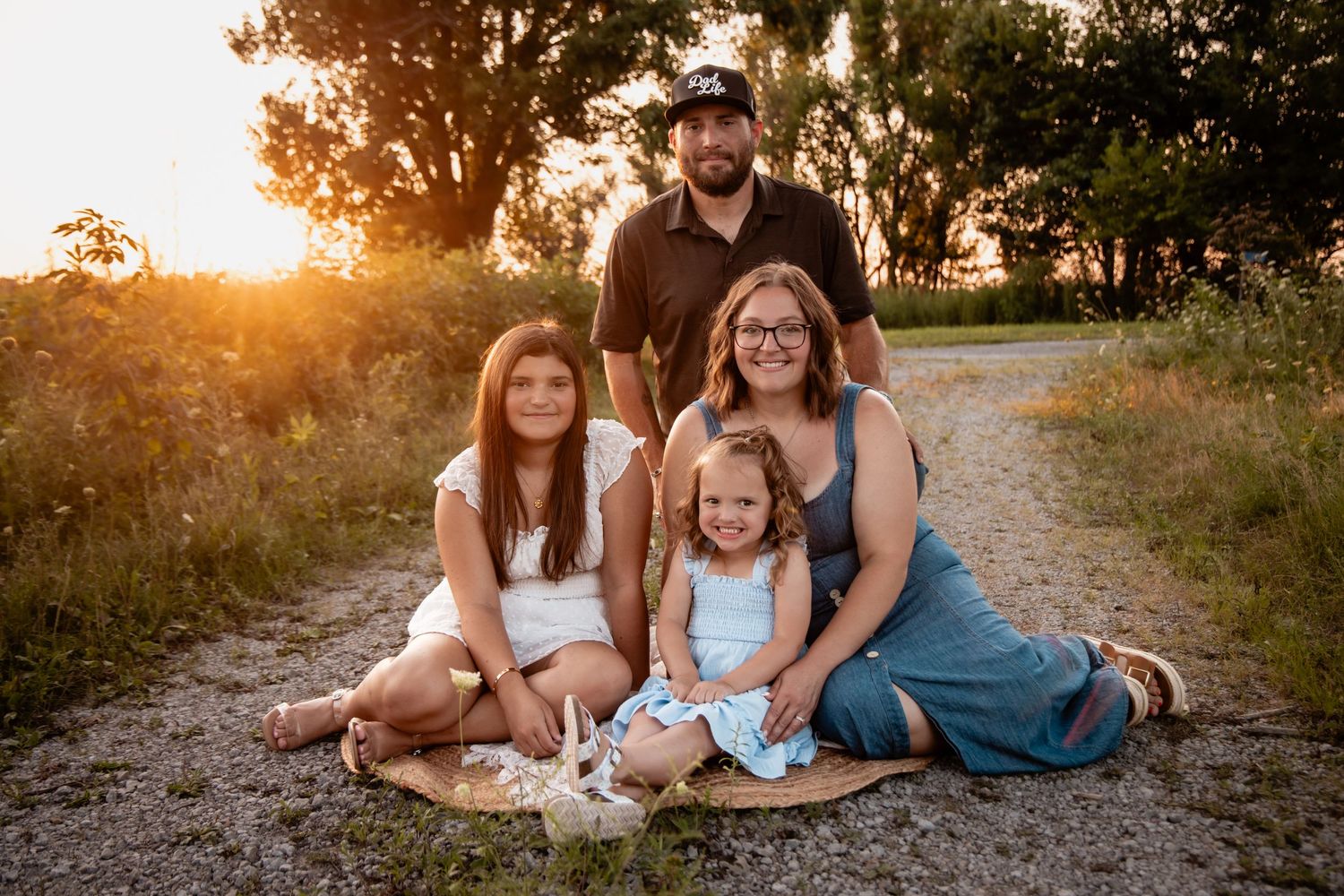 Family of four posing at golden hour on a dirt path surrounded by lush greenery for an outdoor portrait session.