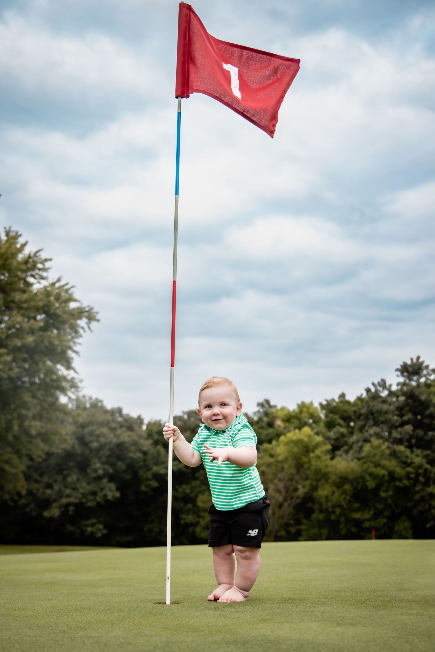 Smiling baby in green striped shirt holds golf flag on putting green, hole 1, with trees and cloudy sky.