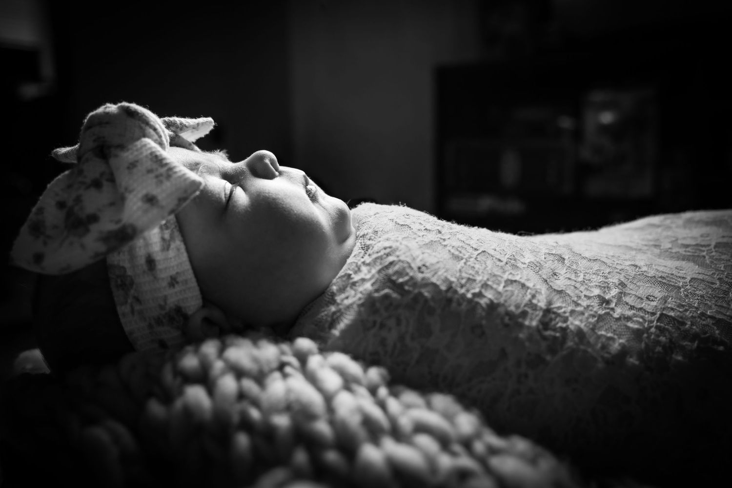 Black and white photo of a newborn baby lying down wearing a floral headband, wrapped in a soft knit blanket.