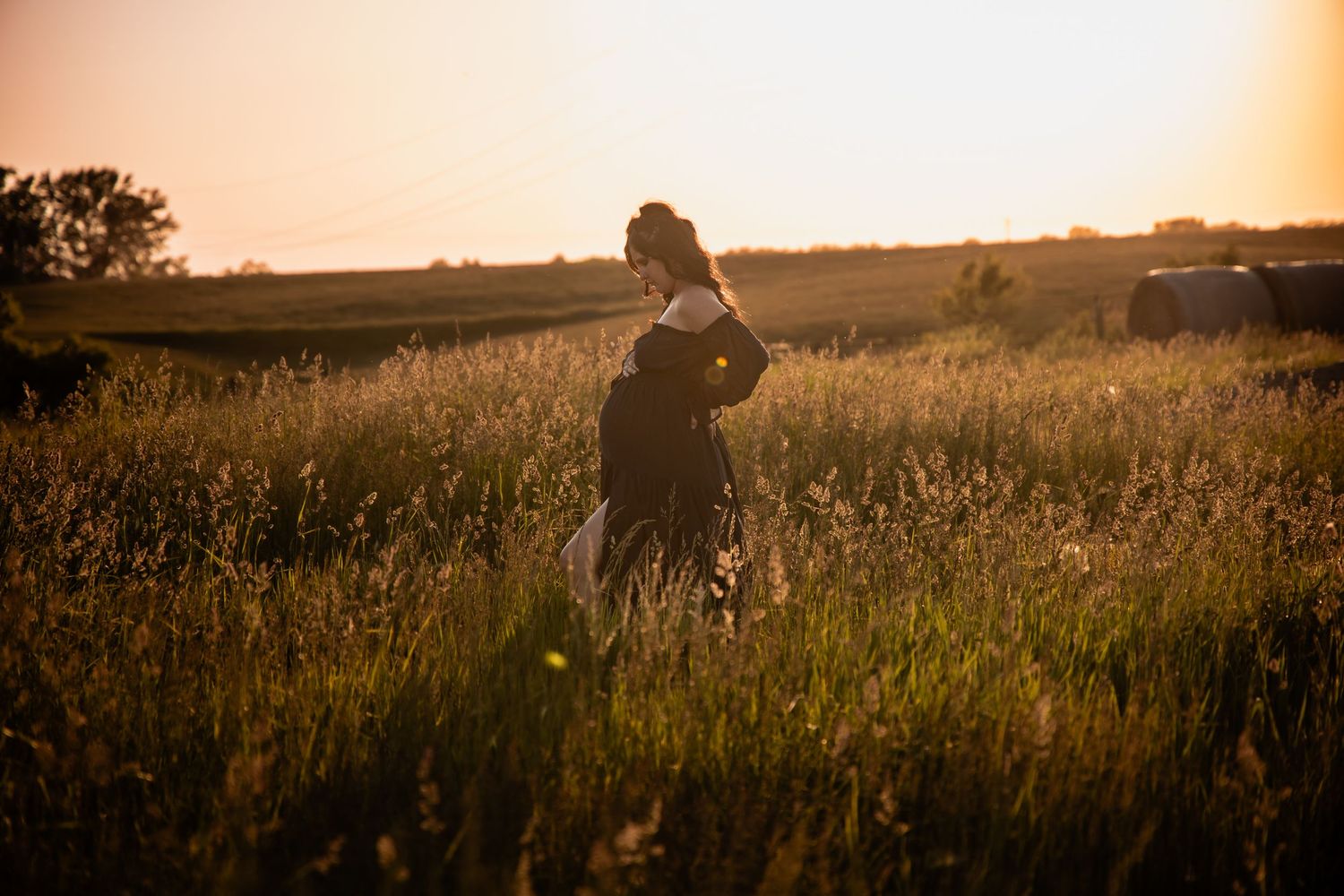 A silhouetted pregnant woman walks through tall golden grass during a stunning sunset.