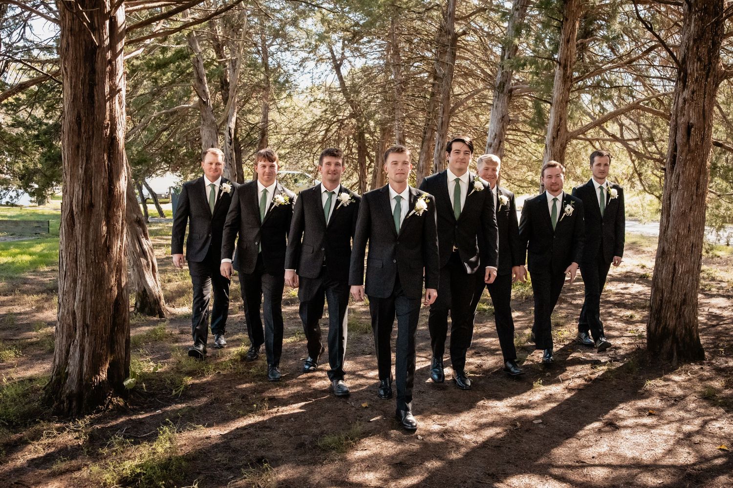 Groomsmen in black suits with green ties walking together through a tree-lined path at an outdoor wedding.
