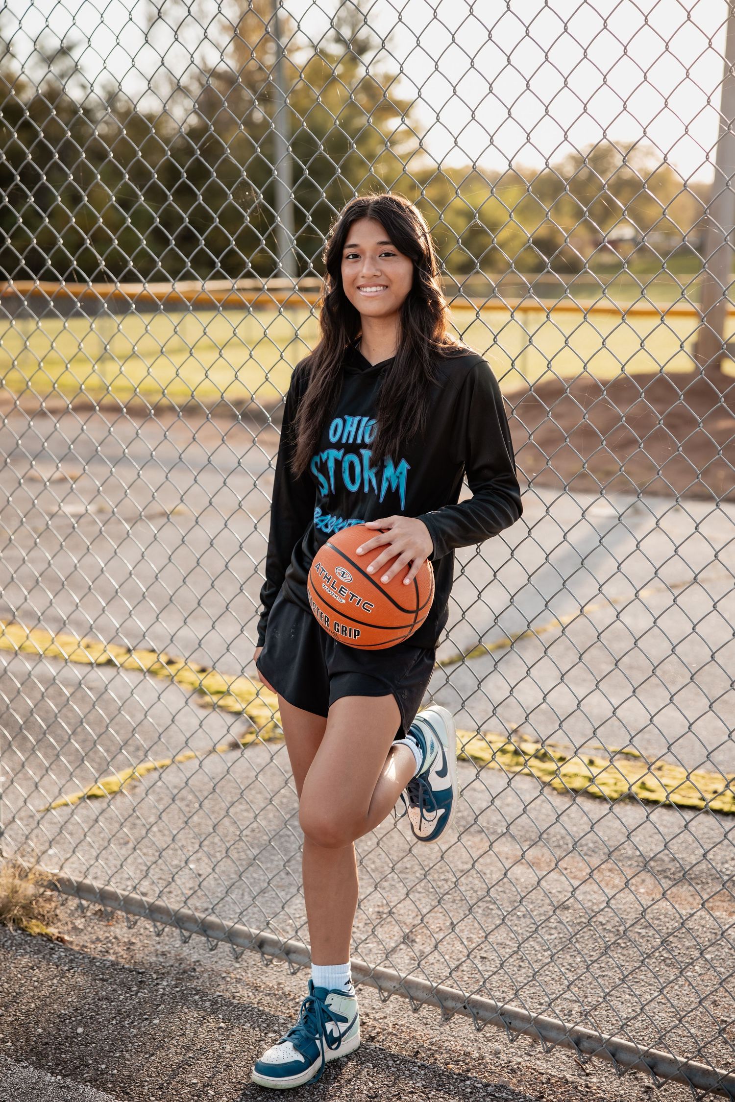 Young girl in Ohio Storm hoodie holds basketball on outdoor court, leaning against chain-link fence, wearing teal sneakers.