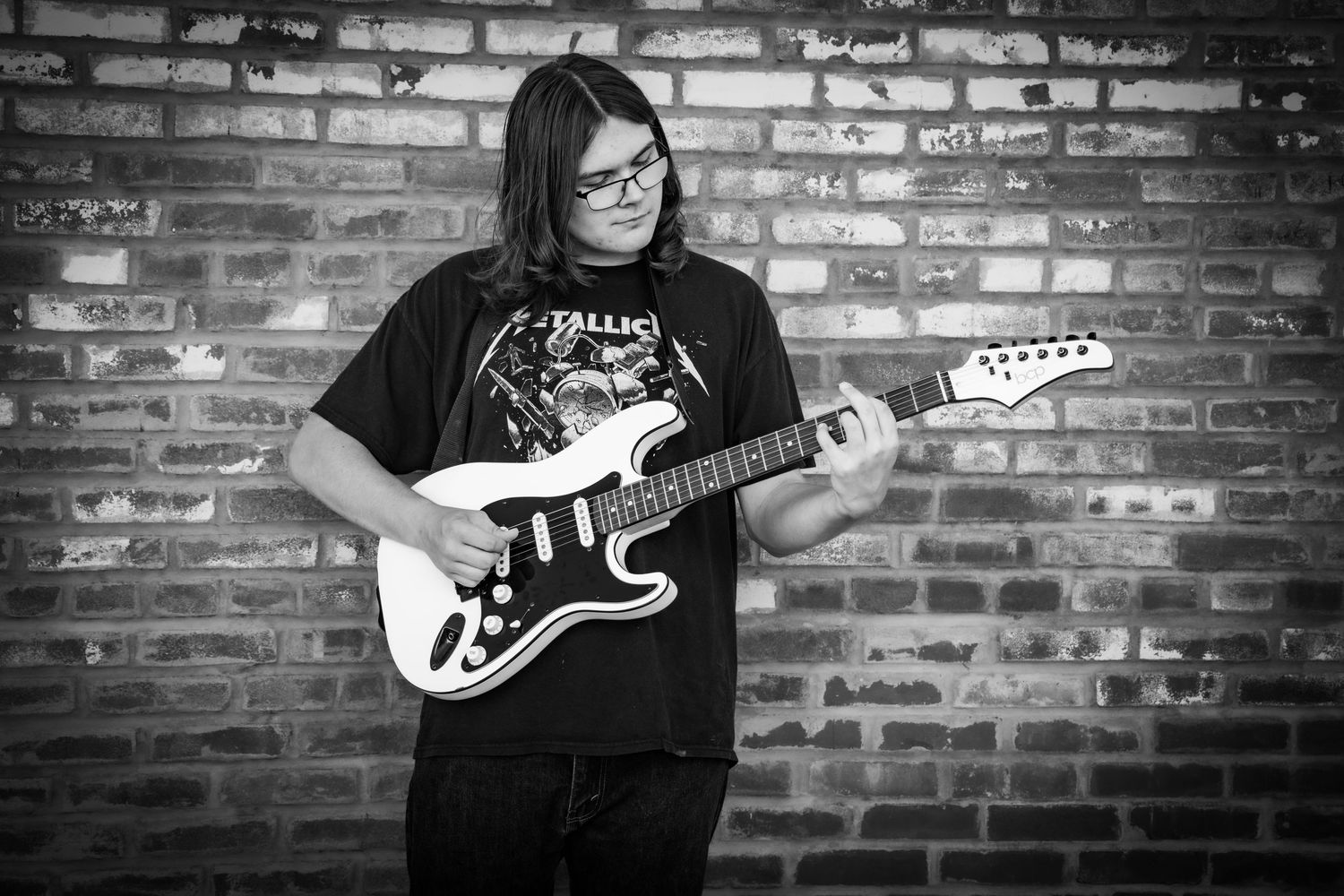 Young guitarist playing electric guitar against brick wall in black and white photo wearing Metallica shirt.
