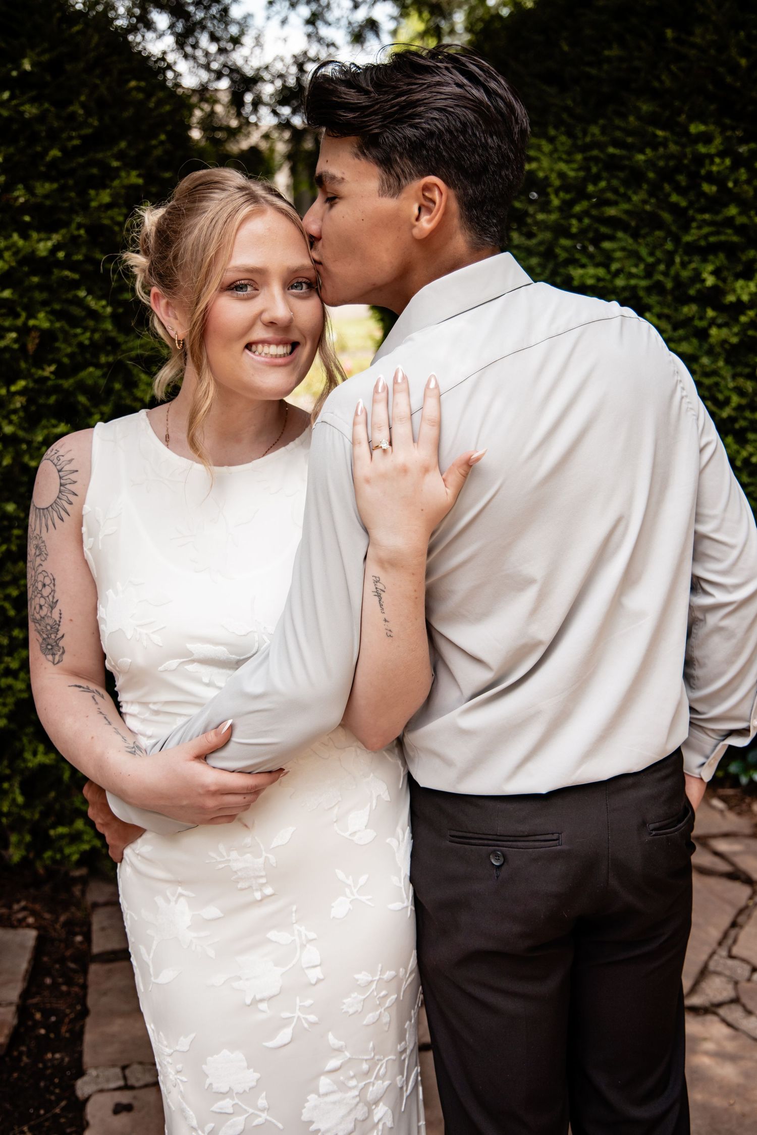 A smiling bride in a white floral gown receives a kiss from her groom in a garden wedding setting.