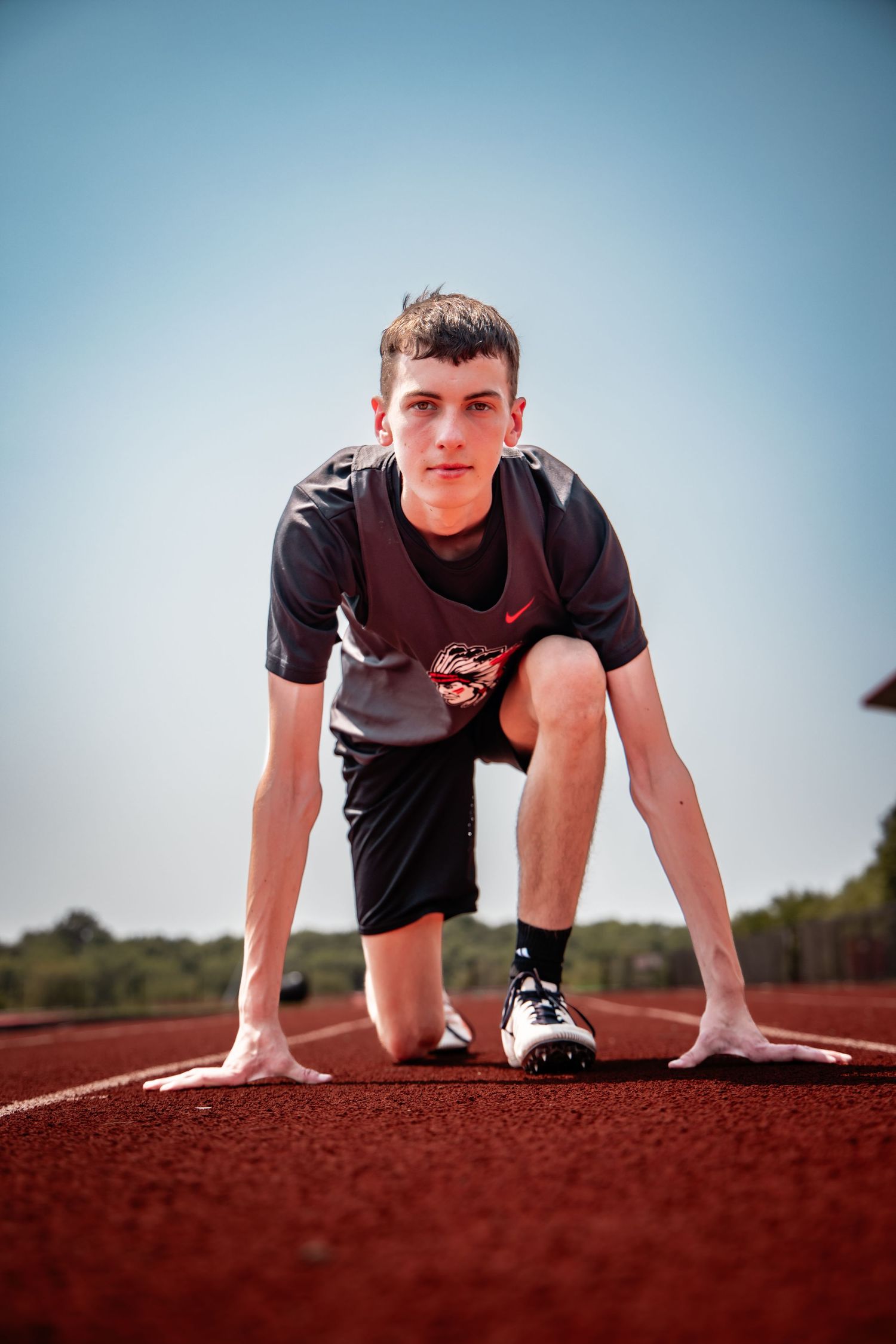 Young male sprinter in starting position on red running track wearing Nike gear, ready to race under clear blue sky.