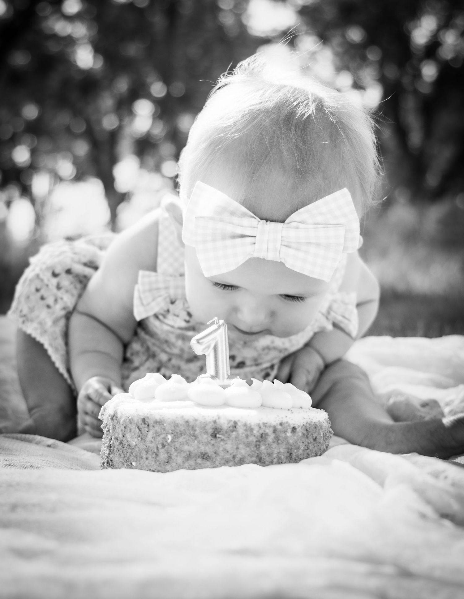 Black and white photo of a baby girl with a bow headband leaning over a small first birthday cake with a number 1 candle.