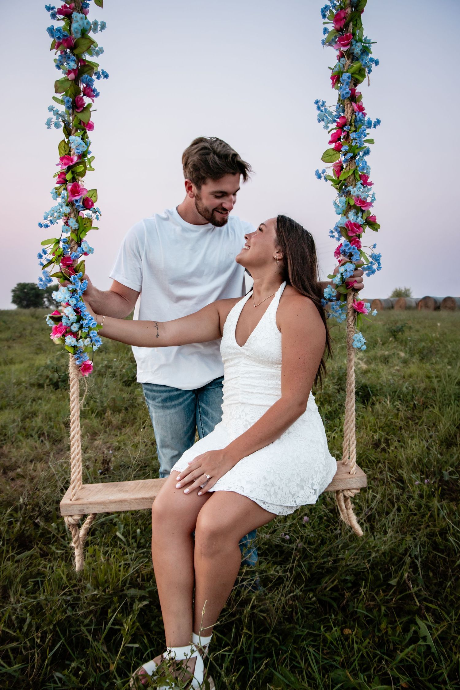 Couple in white outfits sharing a romantic moment on a floral decorated swing in a green field at dusk.