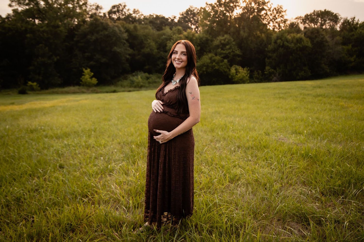 Pregnant woman in brown maxi dress standing in green field at golden hour, cradling baby bump.