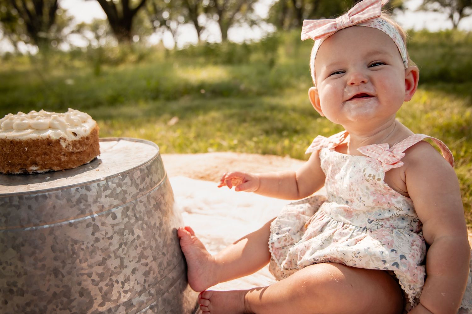 Smiling baby girl in floral dress and pink bow headband sitting outdoors near a small cake during first birthday celebration.