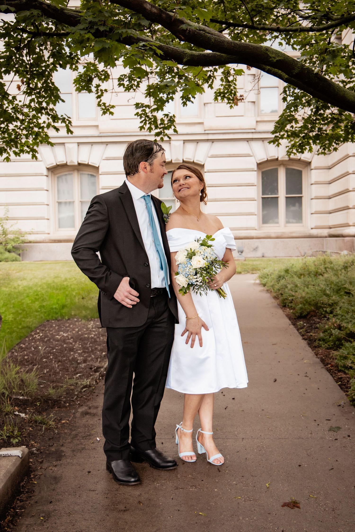 Bride in white off-shoulder dress holds bouquet as groom in dark suit kisses her cheek on garden path.