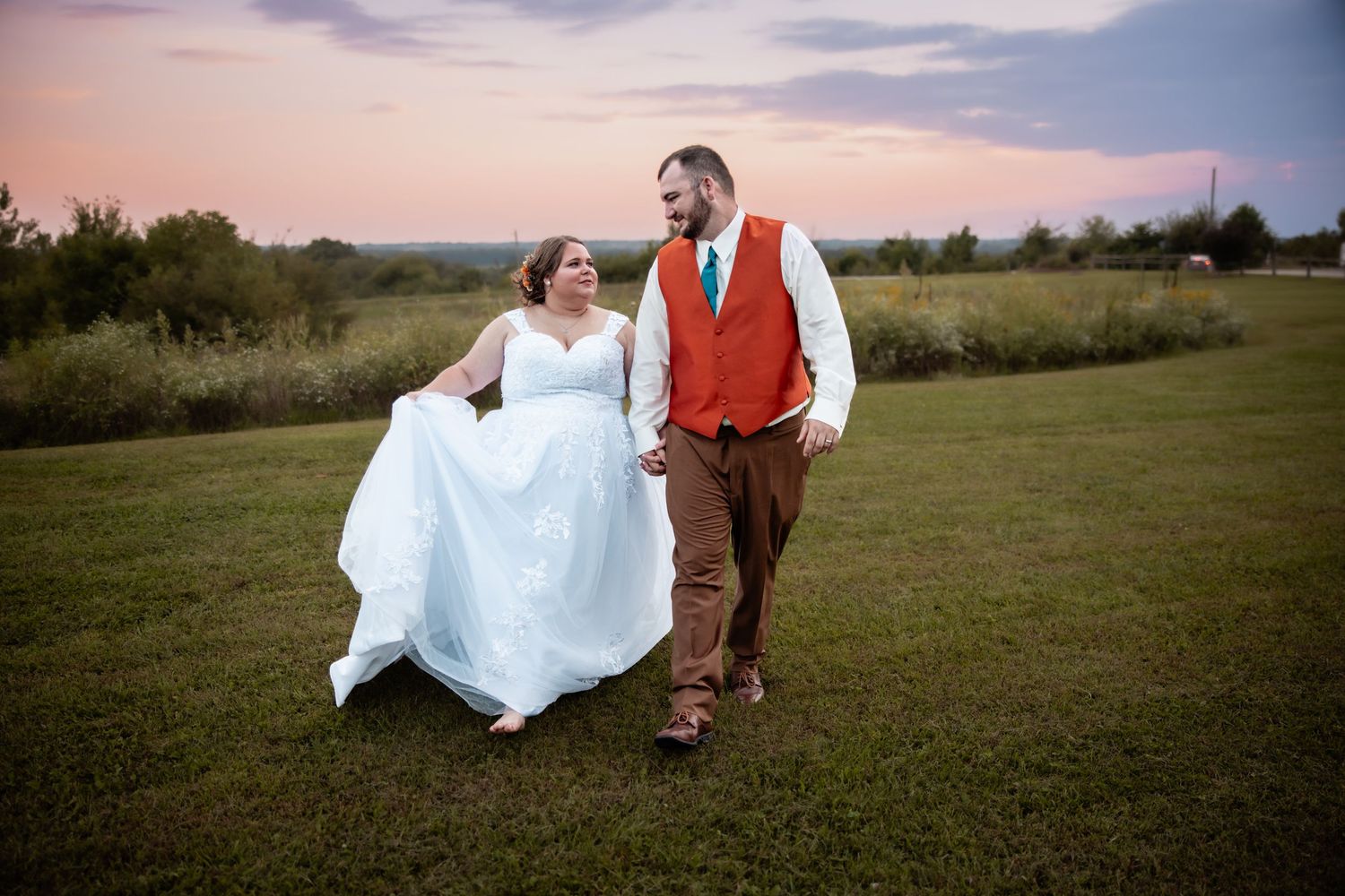 Bride in white gown and groom in rust vest walk hand-in-hand across green field at sunset.