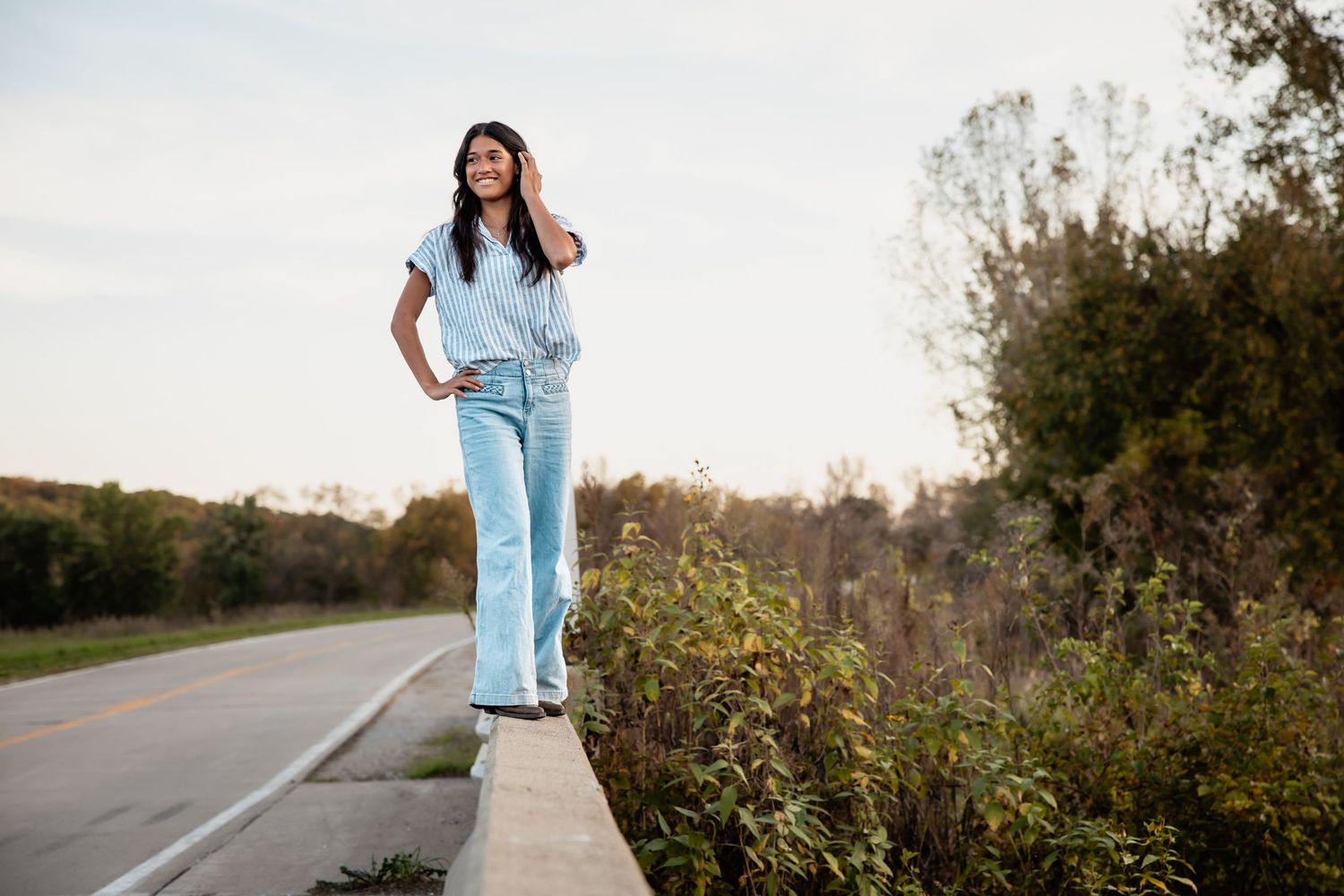Young woman in light blue denim outfit standing on a concrete ledge beside a road, smiling outdoors.