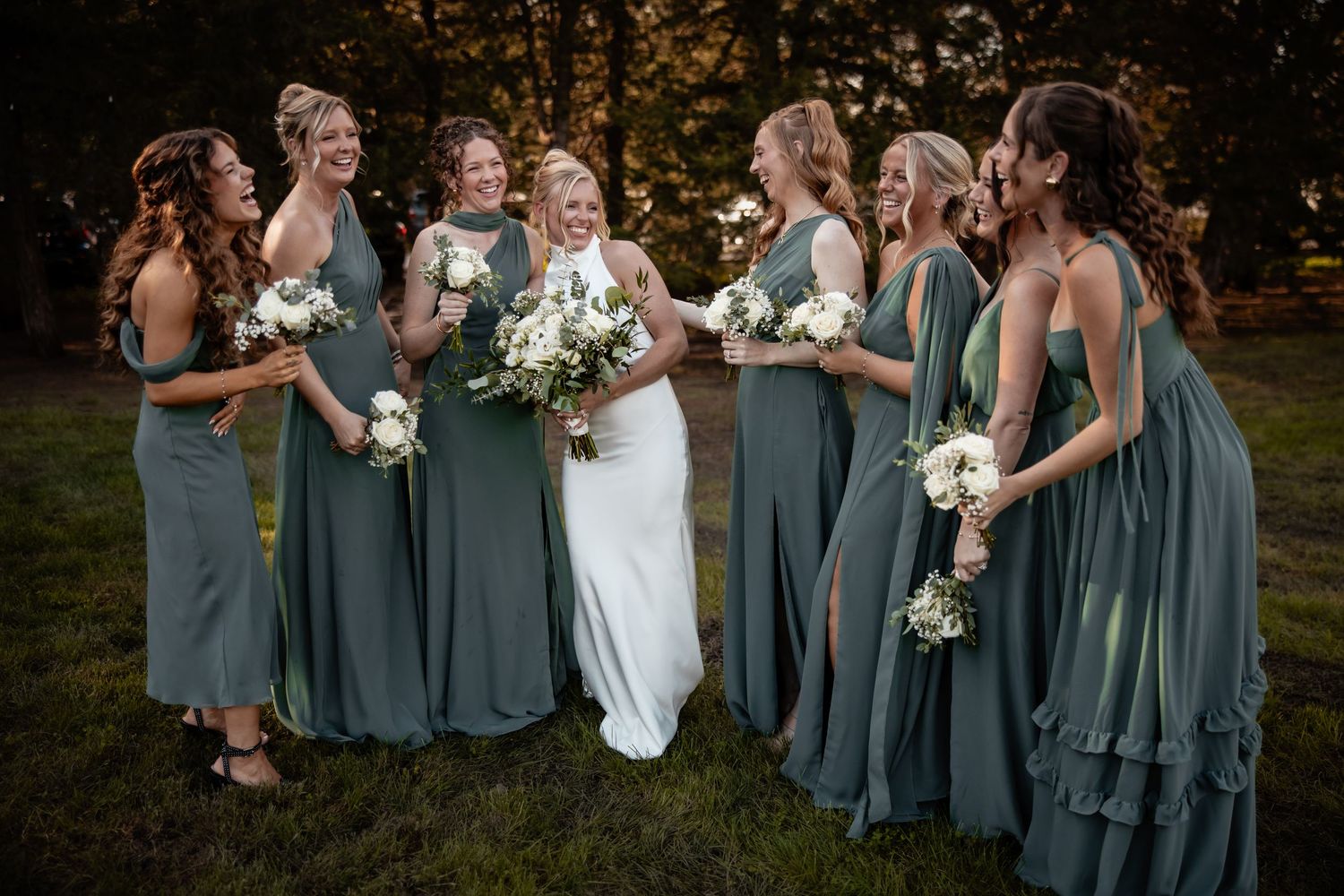 Bride in white gown with six bridesmaids in sage green dresses holding white bouquets, laughing outdoors.