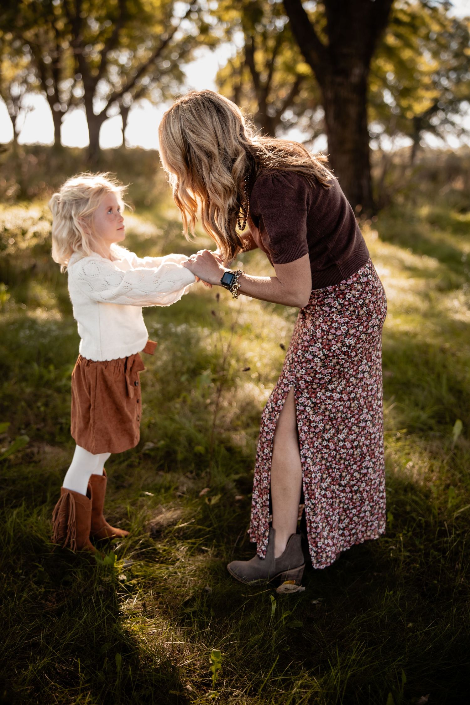 A mother and young daughter hold hands in a sunlit autumn field, both wearing rustic boho outfits with skirts and boots.