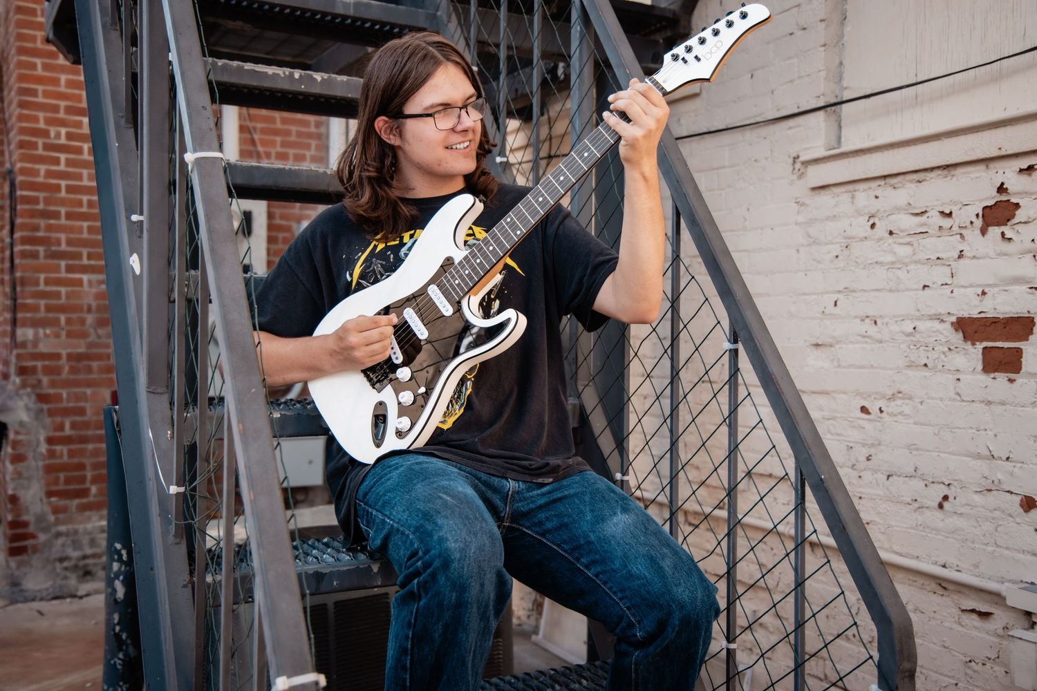 Young guitarist with long hair and glasses playing white electric guitar on metal stairs outdoors.
