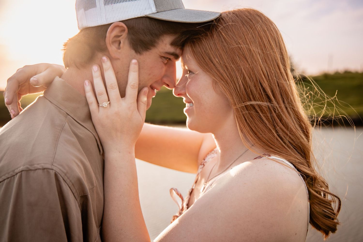 An engaged couple leans in close with foreheads nearly touching, engagement ring visible, bathed in warm sunset light.