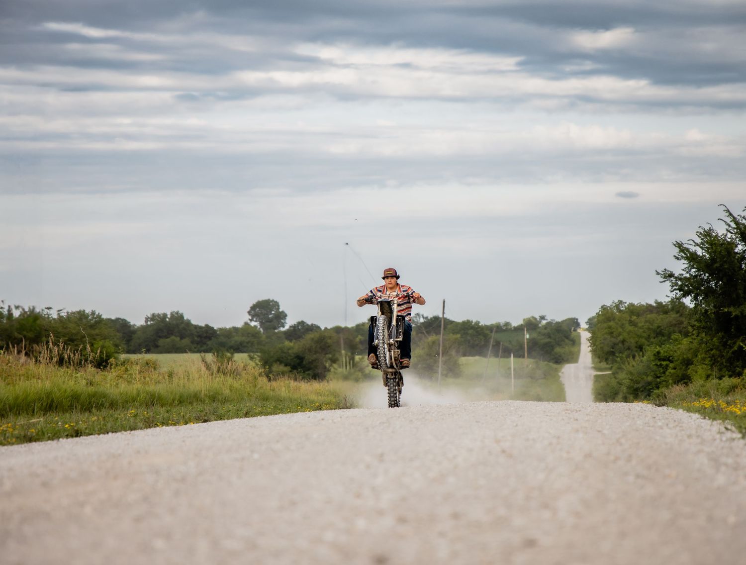 Motorcyclist performing a wheelie on a dusty rural road surrounded by green fields under a cloudy sky.