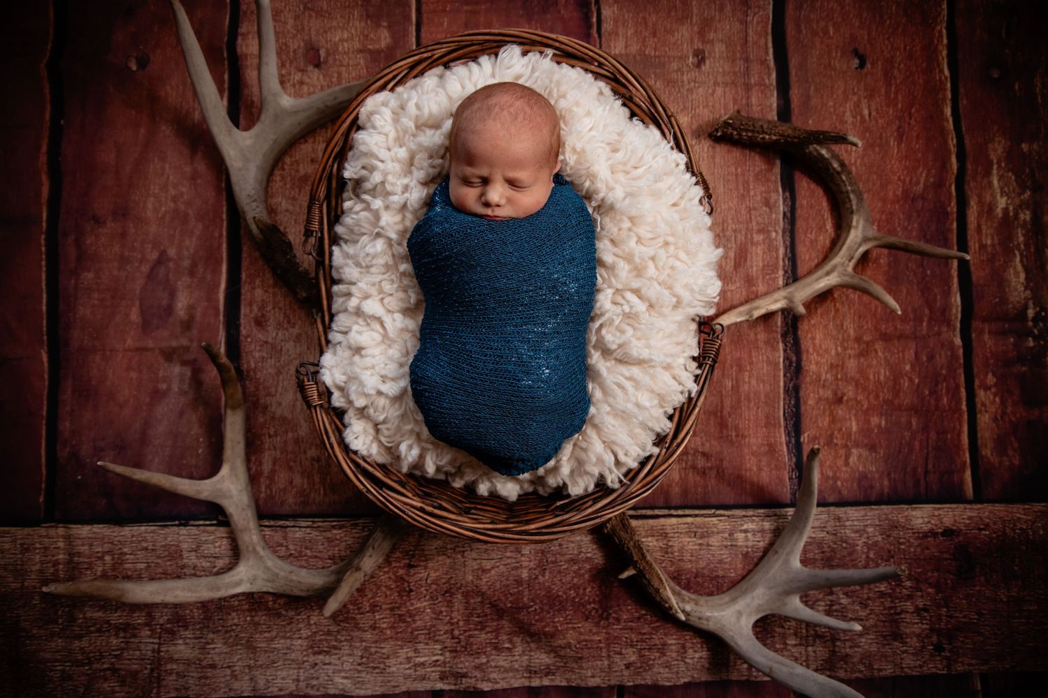 Newborn baby swaddled in blue wrap, nestled in a wicker basket with white fur, surrounded by deer antlers on rustic wood.