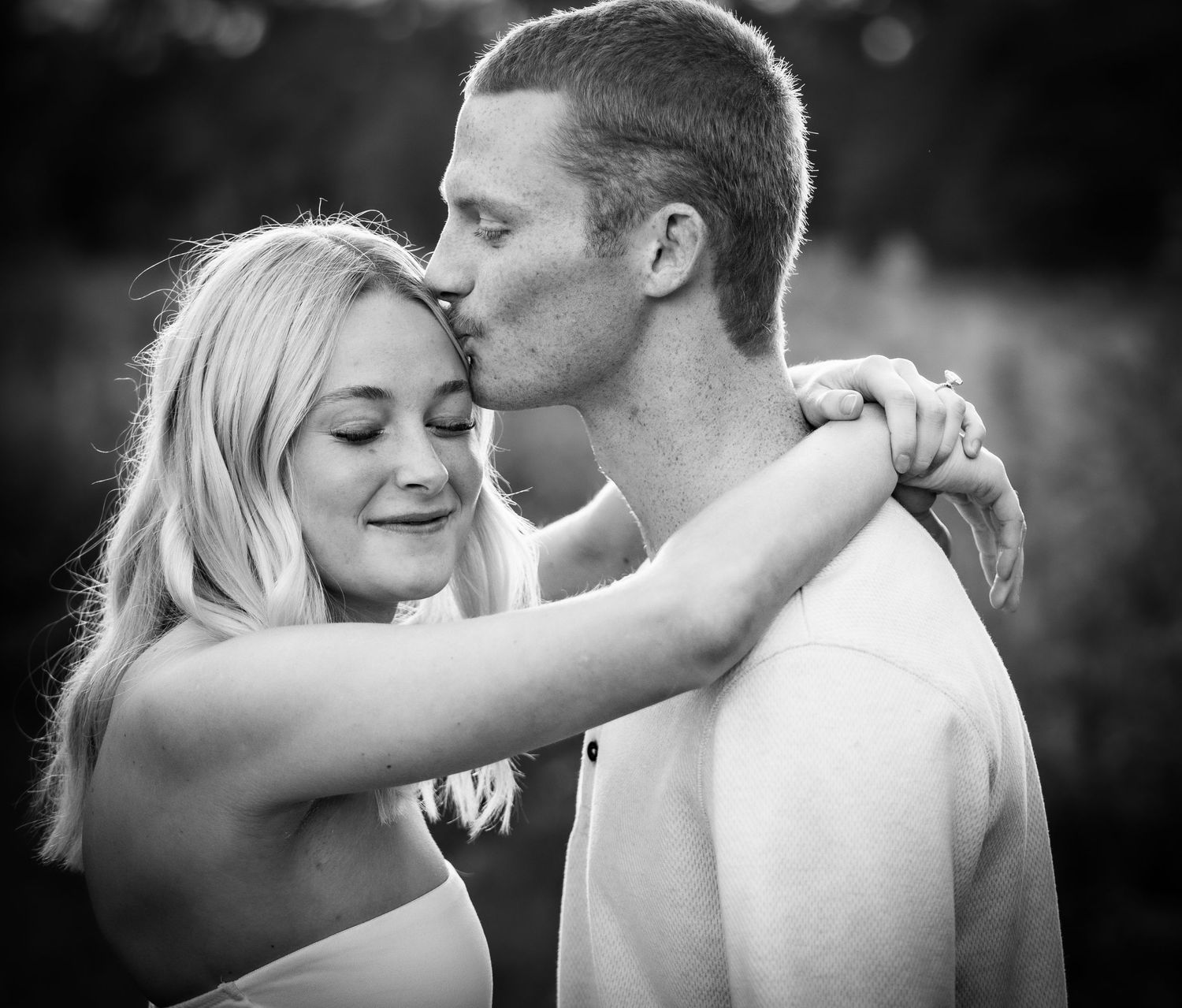Black and white photo of a young couple embracing outdoors, man kissing woman on the forehead.
