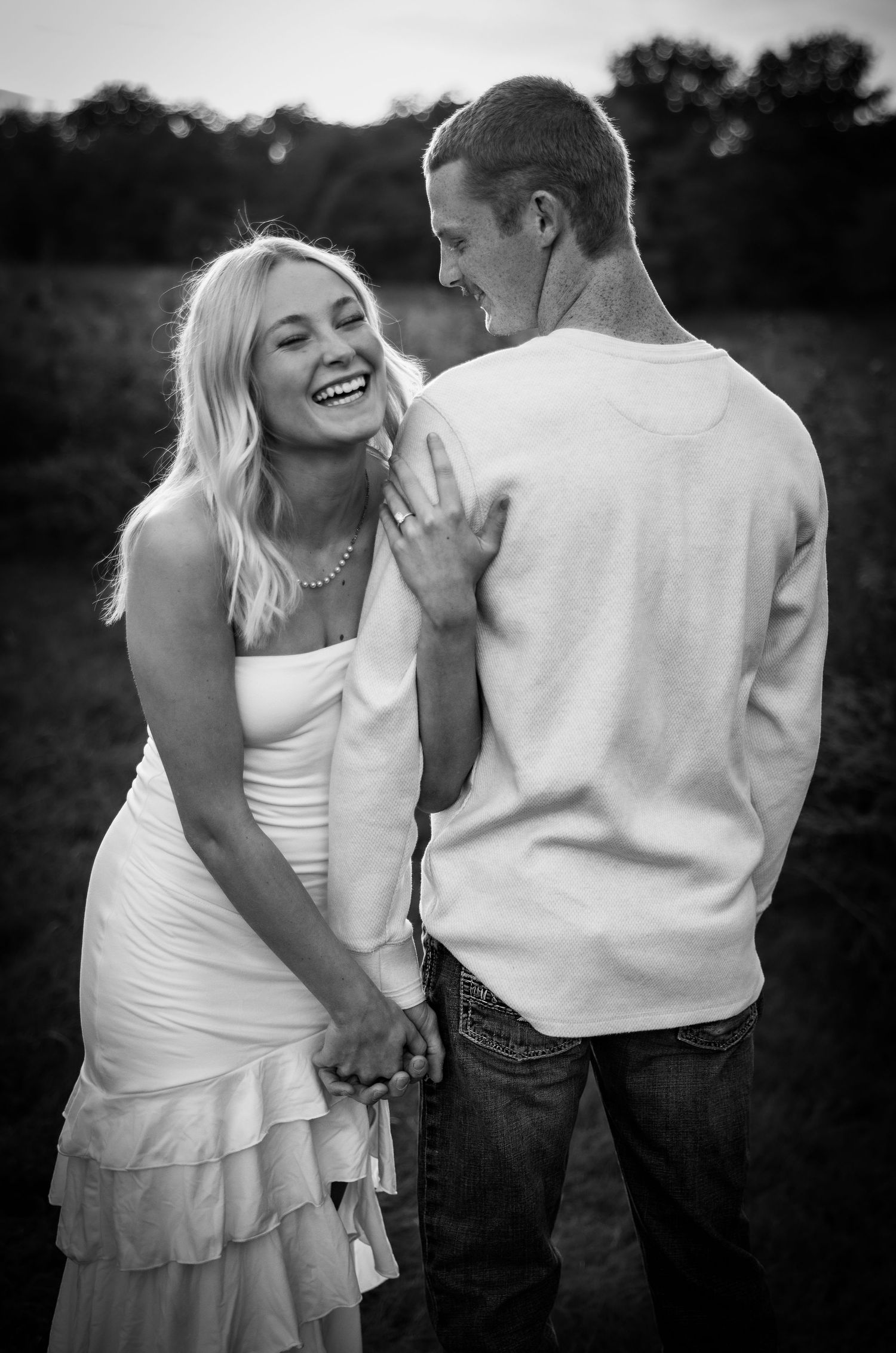 A joyful couple laughs together outdoors, woman in white ruffled dress holding hands with man in sweatshirt and jeans.