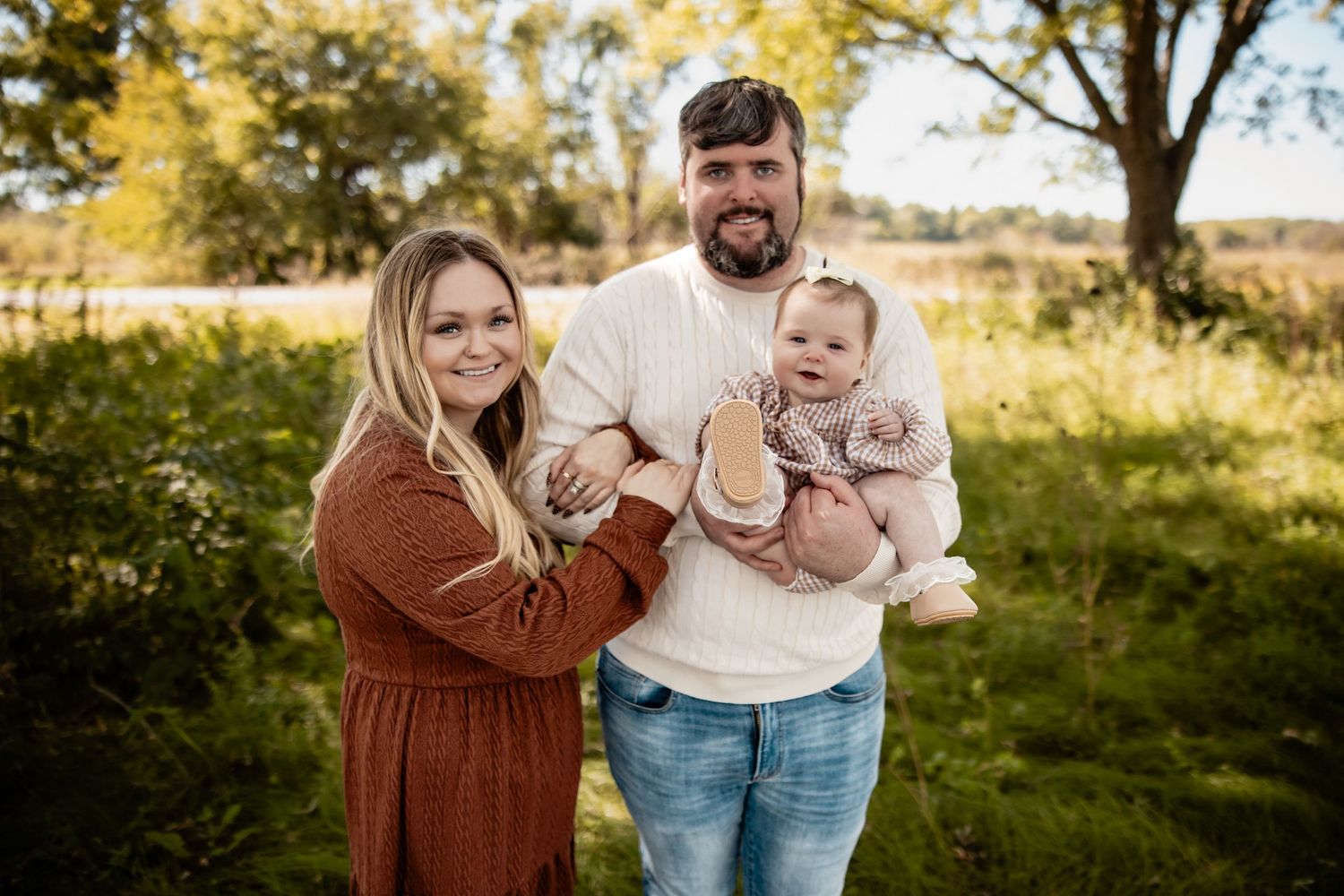 A young family poses outdoors in fall, man holding baby girl, woman in rust dress, green trees behind them.
