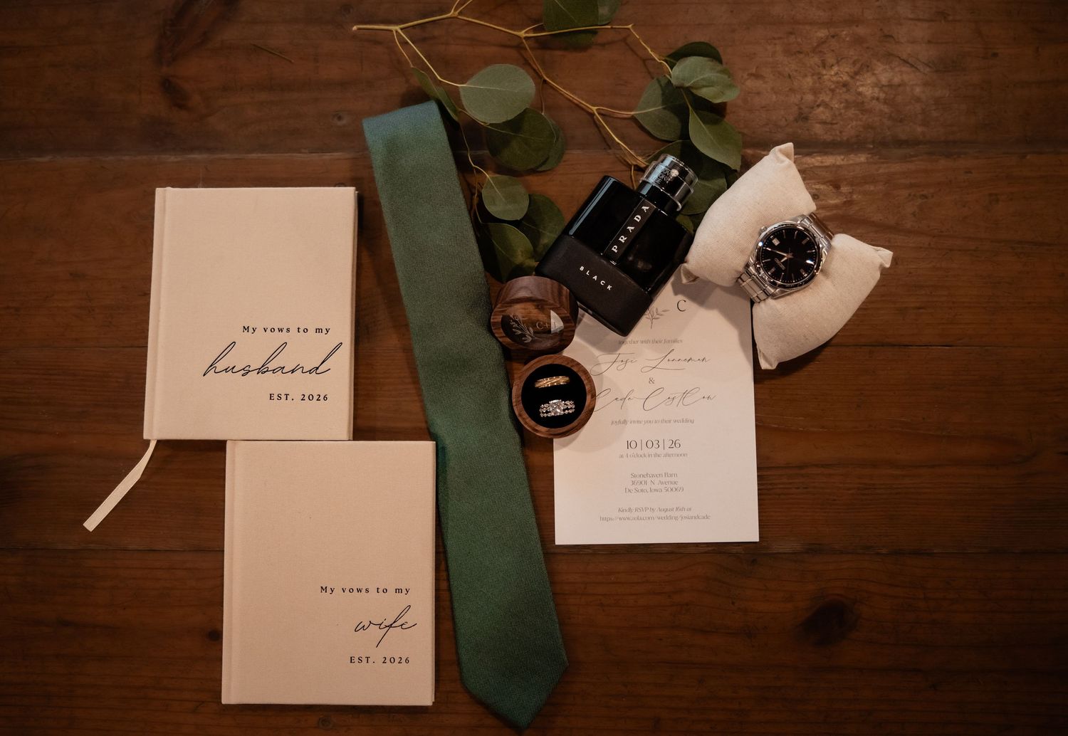 Wedding flat lay with vow books, green tie, watch, ring box, cologne, and eucalyptus on rustic wood.