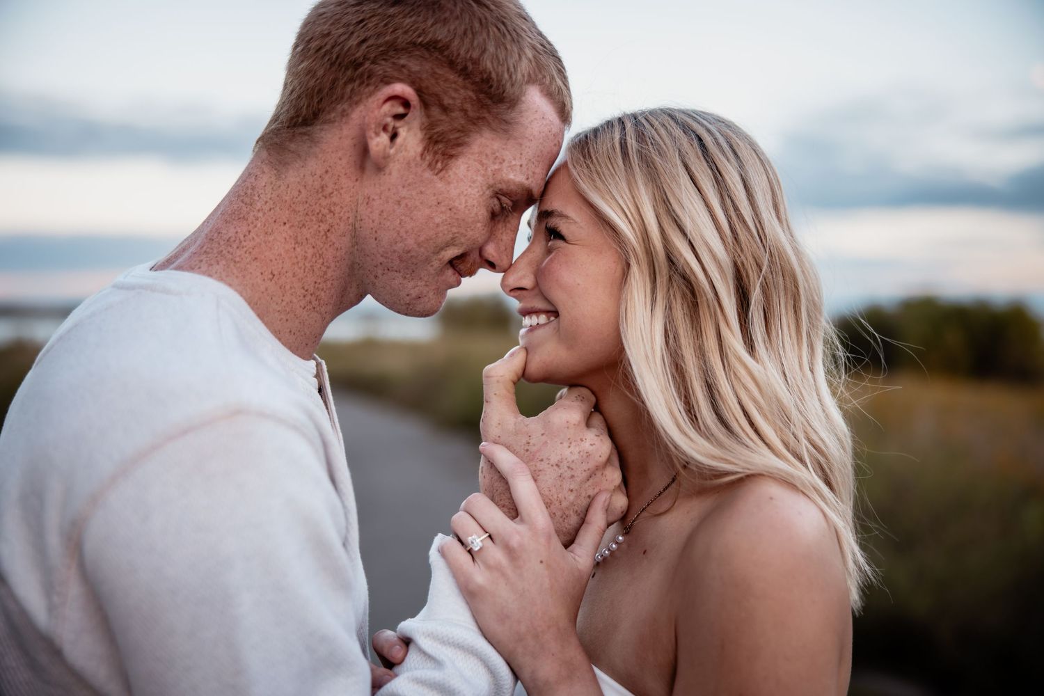A smiling couple with foreheads touching, sharing a romantic moment outdoors at dusk, woman wearing an engagement ring.