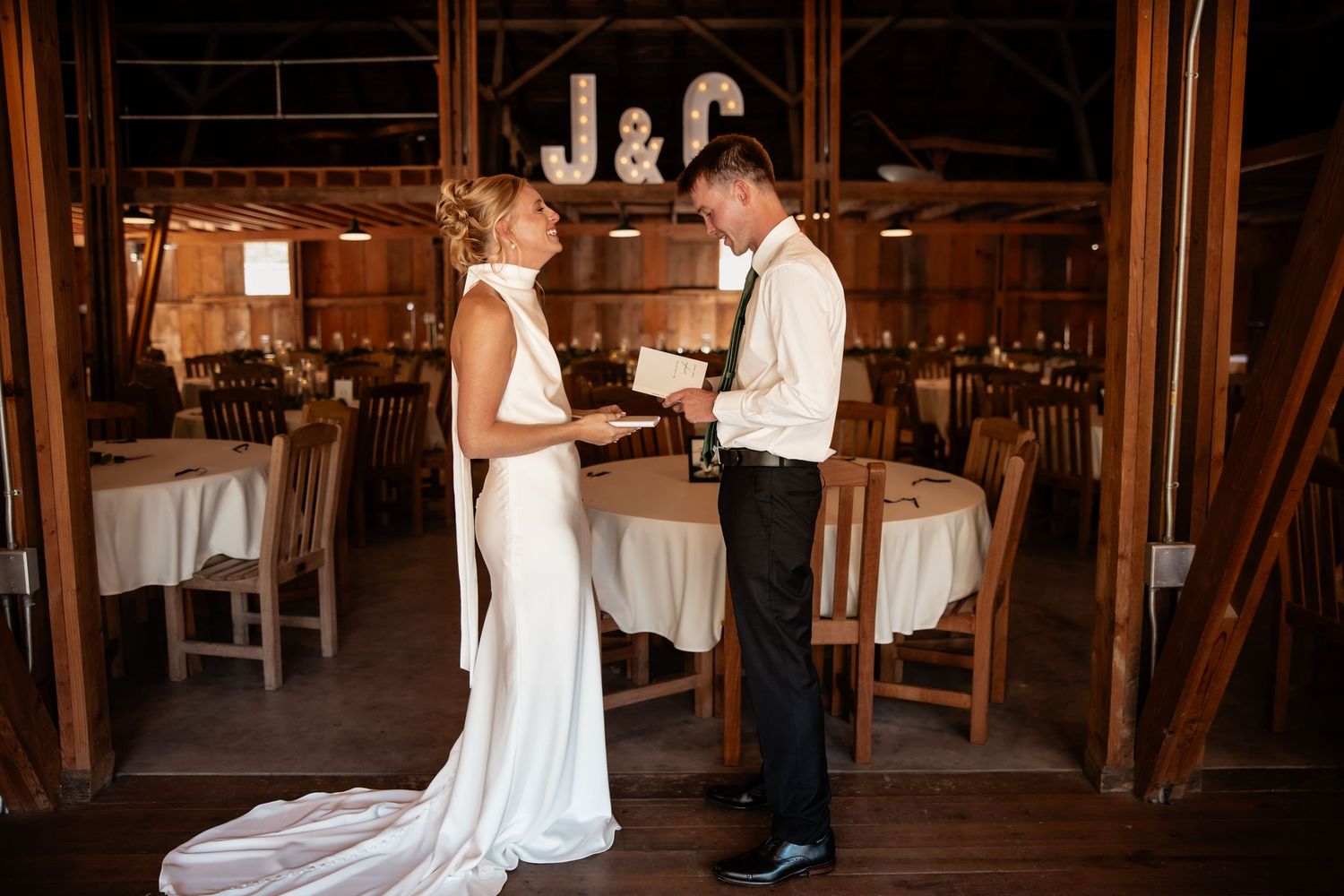Bride and groom exchanging vows inside a rustic barn wedding venue with J&C monogram lights in background.
