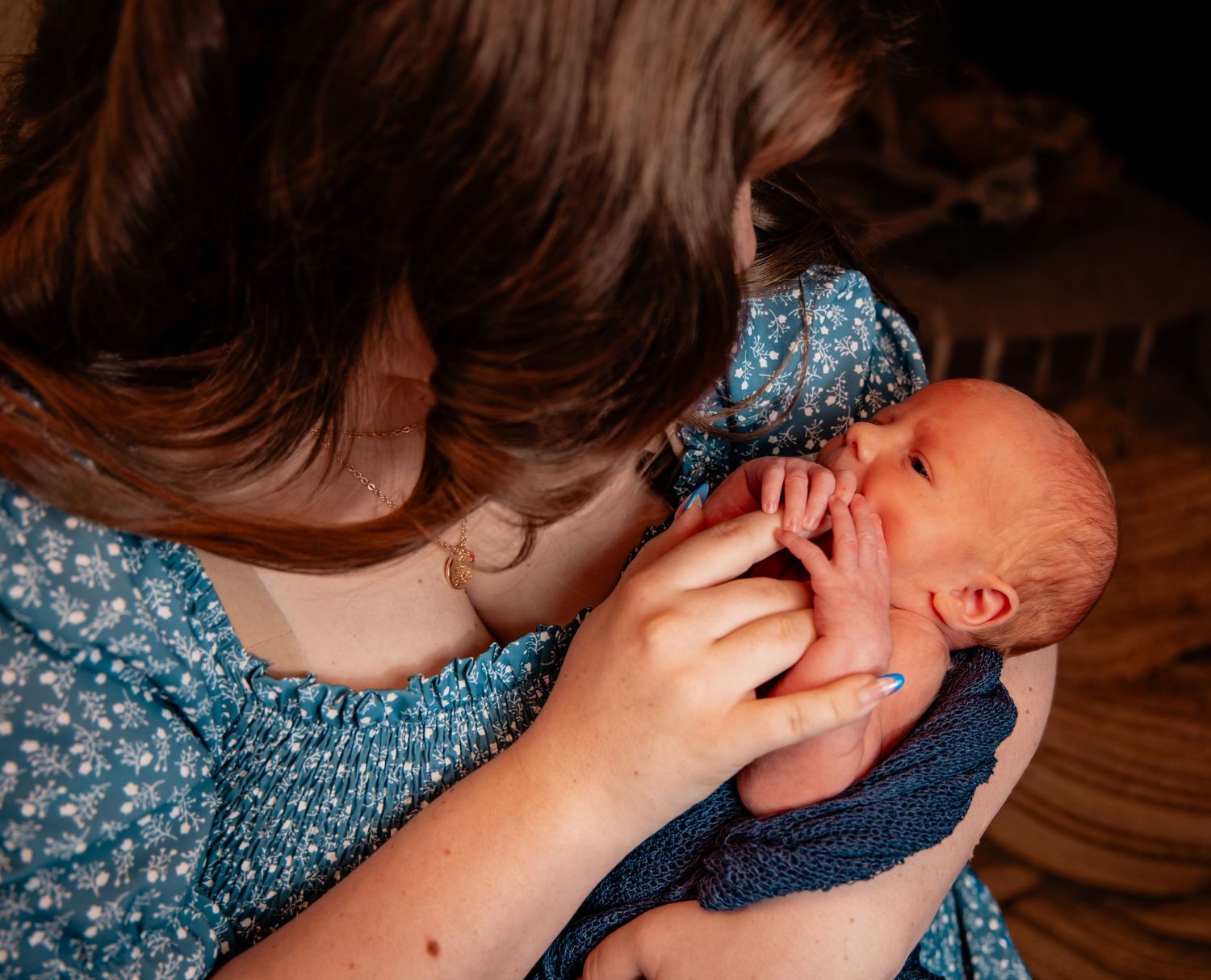 A young girl in a blue floral dress tenderly holds and gazes at a sleeping newborn baby wrapped in a navy blanket.