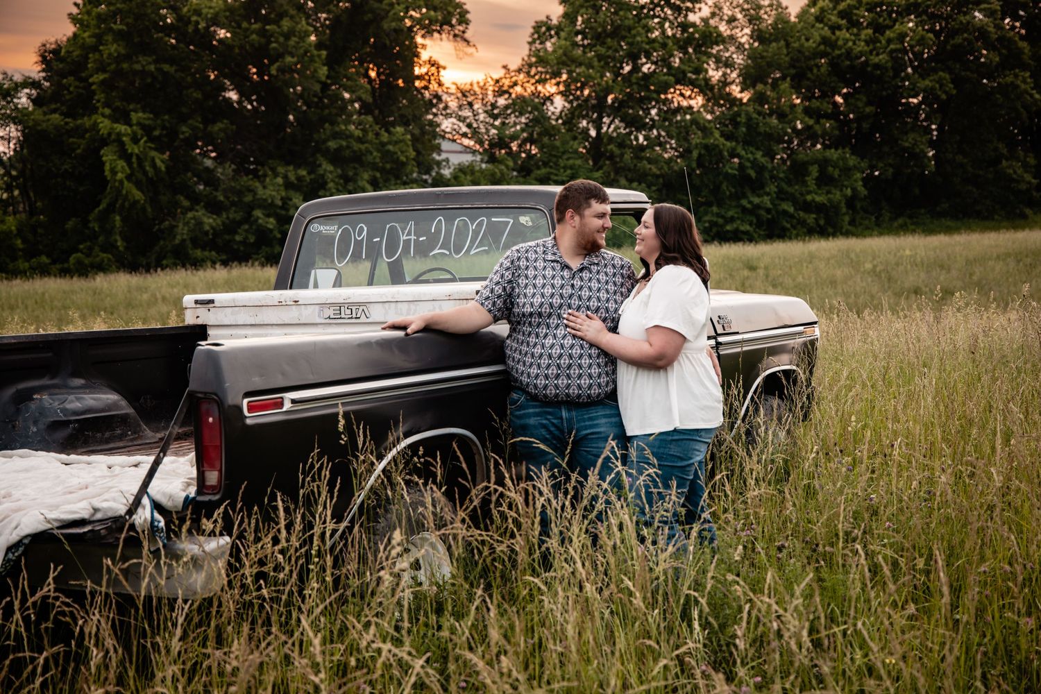 Engaged couple leaning against vintage truck in tall grass field at sunset.