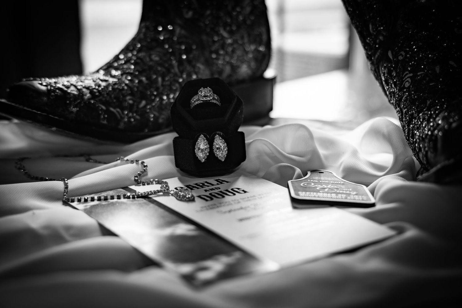 Black and white flat lay of wedding details including diamond ring, earrings, boots, and invitation on white fabric.