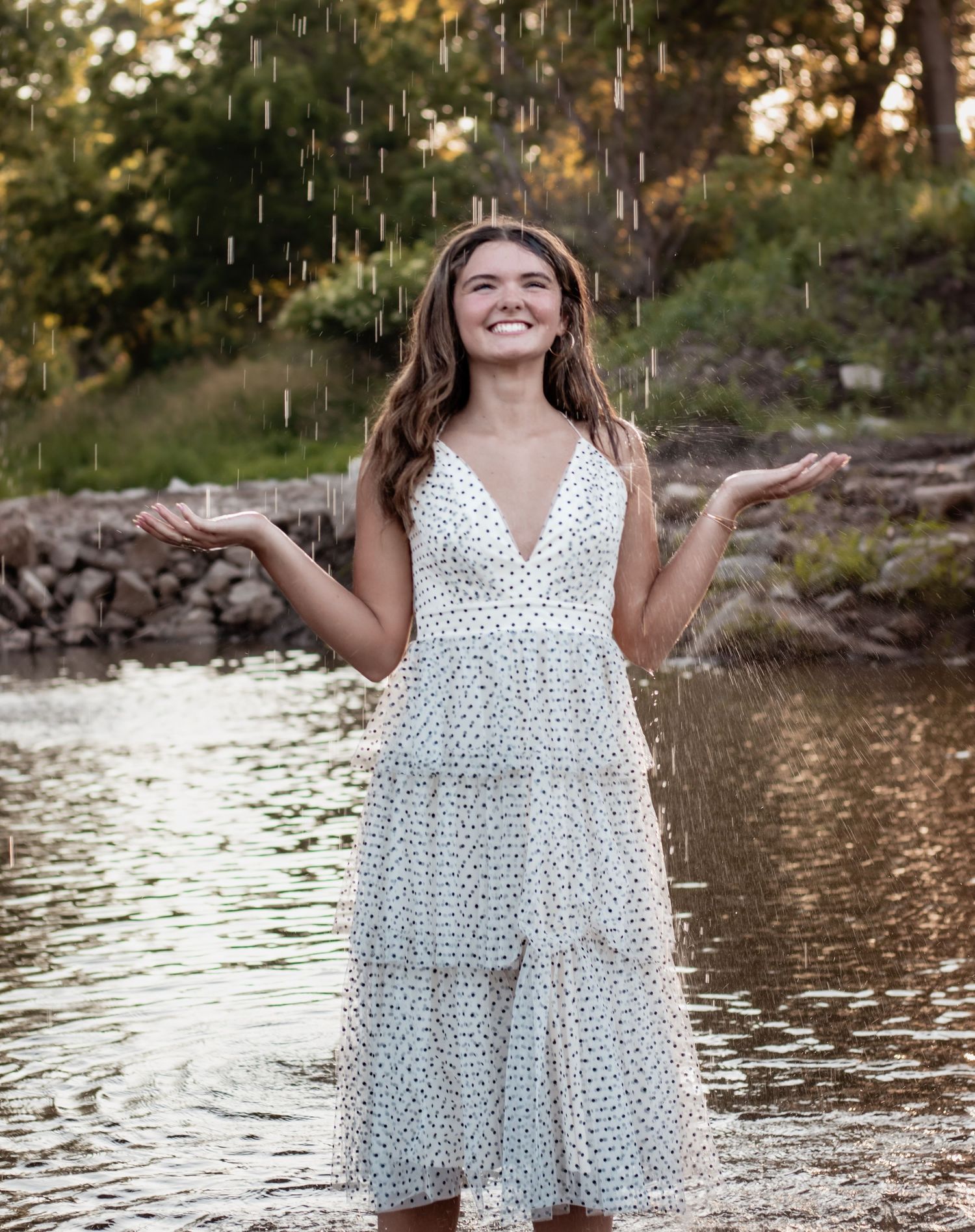 Smiling young woman in white polka dot dress standing in water with hands out, enjoying rain in a scenic outdoor setting.