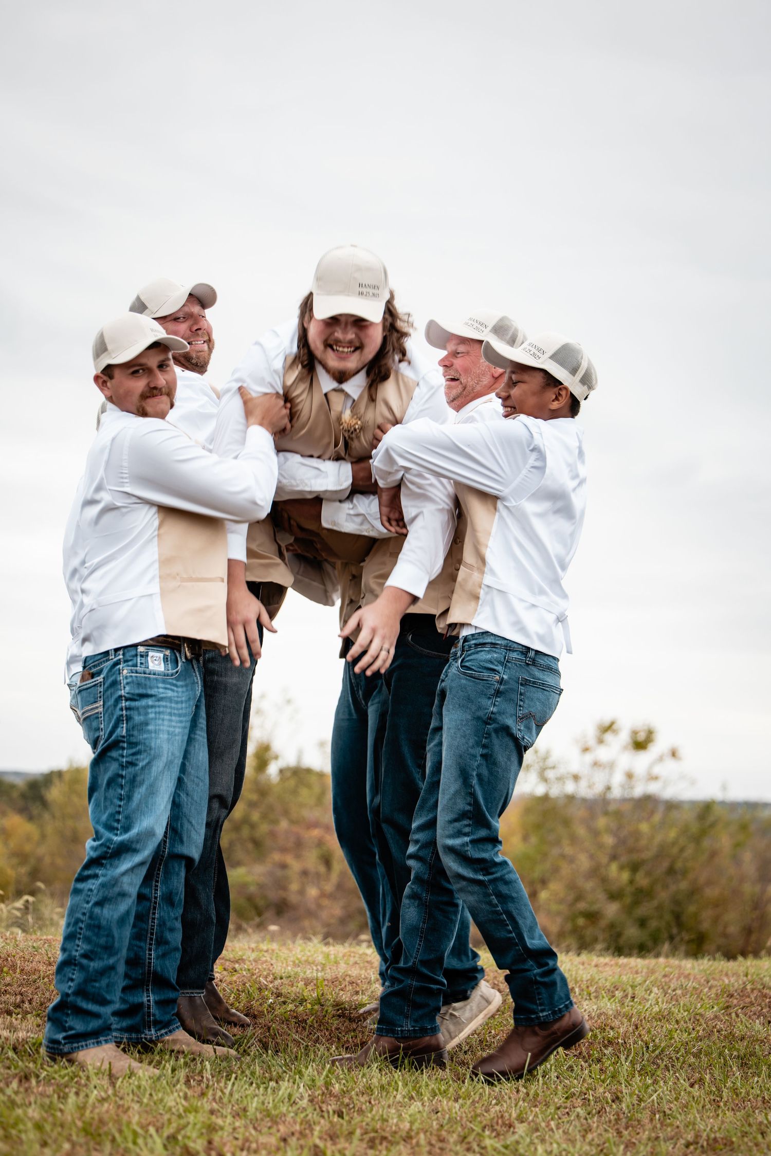 Groom in white shirt and cap being lifted by laughing groomsmen in a field outdoors.