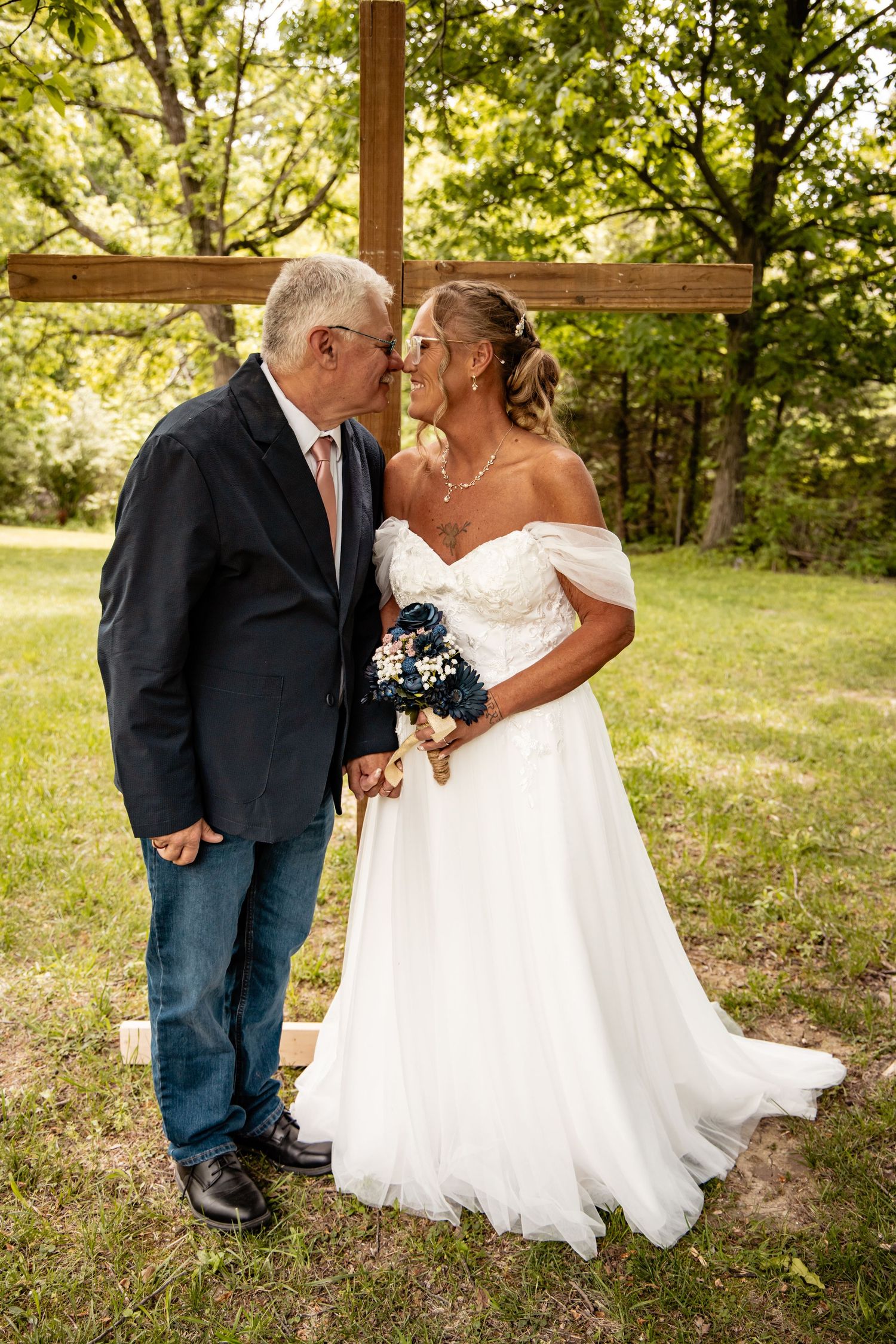 Bride and groom share a romantic nose-to-nose moment in front of a wooden cross outdoors.