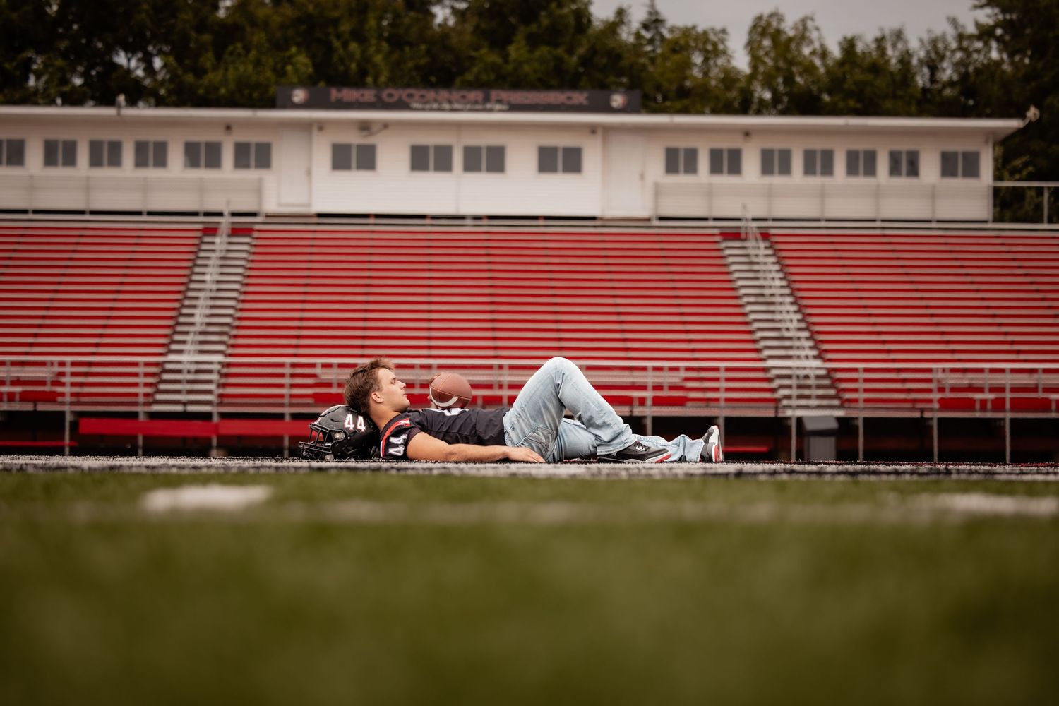 Athlete in football jersey relaxing on track beside red stadium bleachers on a cloudy day.