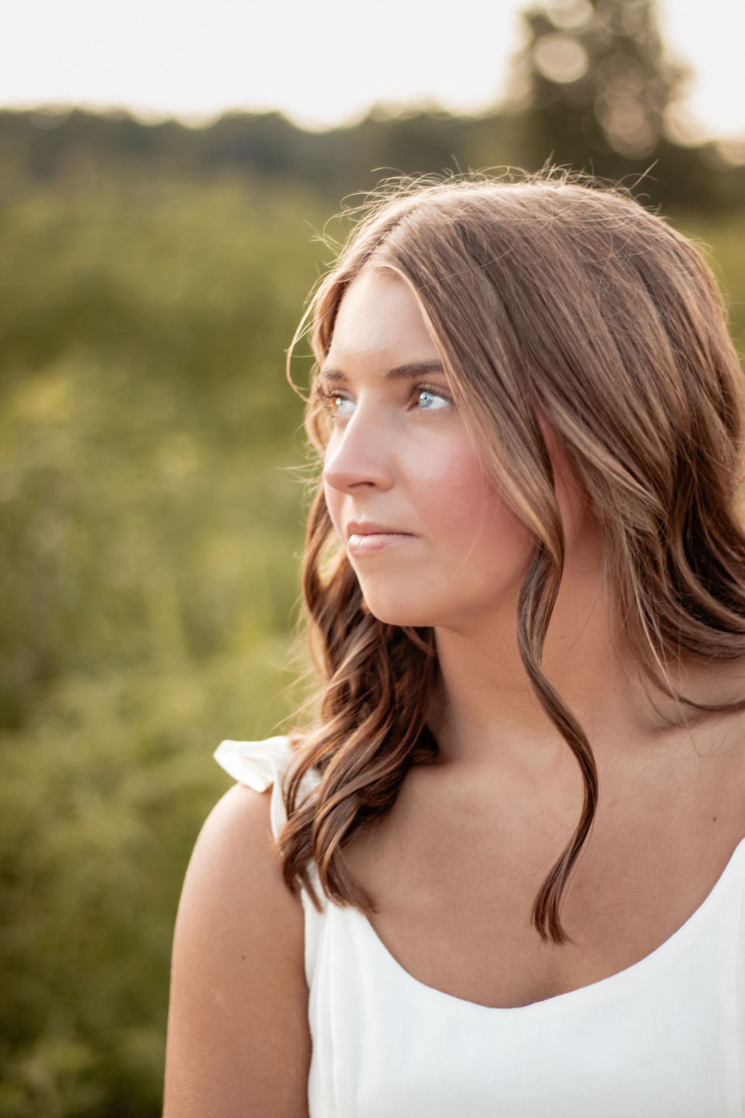 Young woman with wavy brown hair in white dress gazing away in golden hour outdoor portrait.