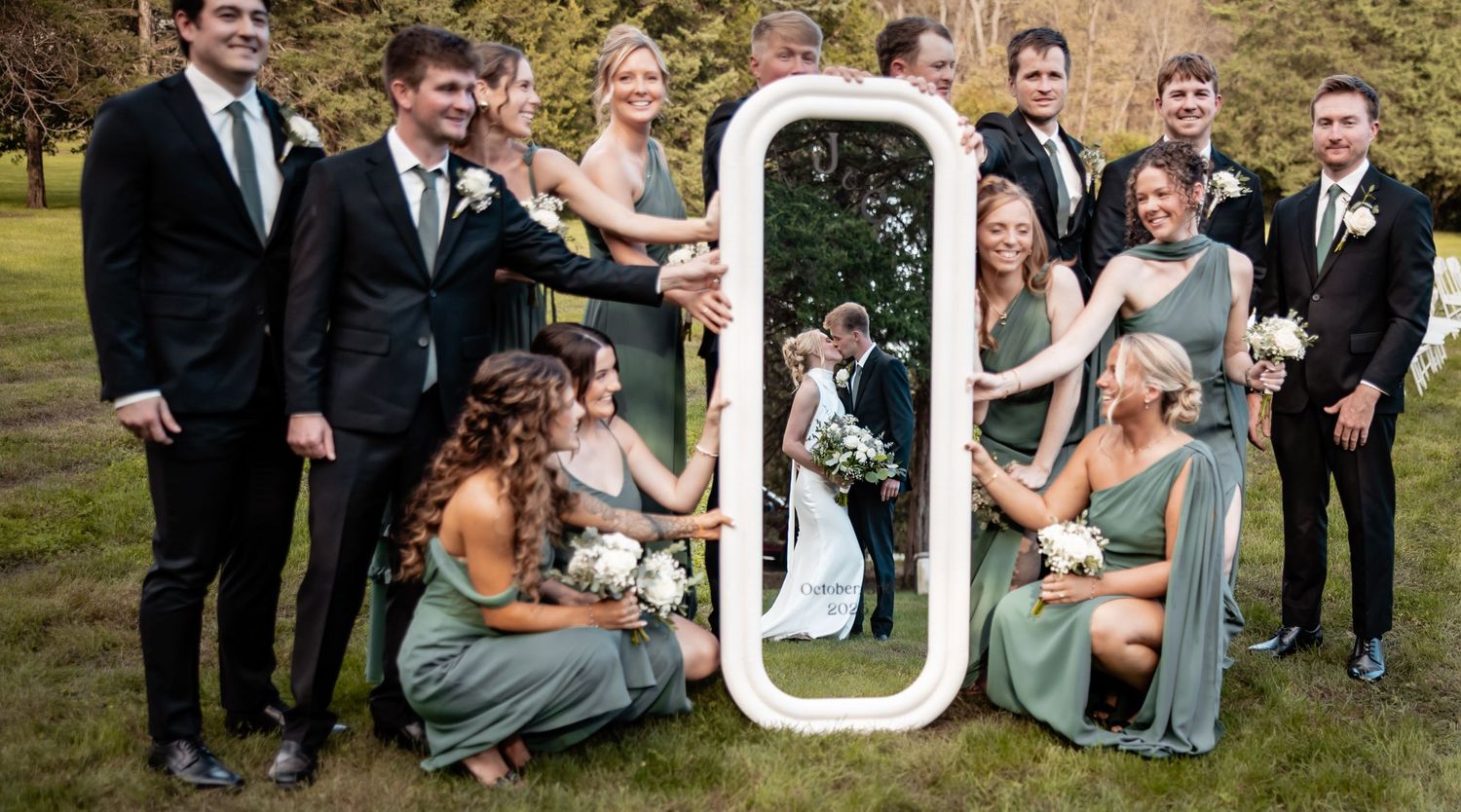 Wedding party poses outdoors with a decorative white mirror reflecting the bride and groom kissing, bridesmaids in sage green gowns.