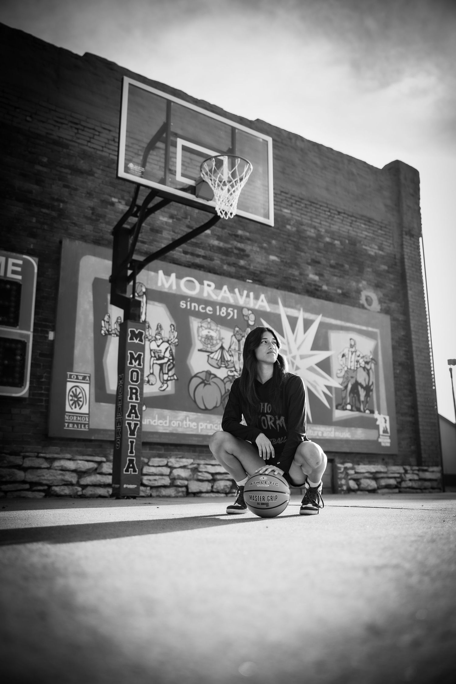 Black and white photo of a young woman crouching with a basketball on an outdoor court near a Moravia mural.