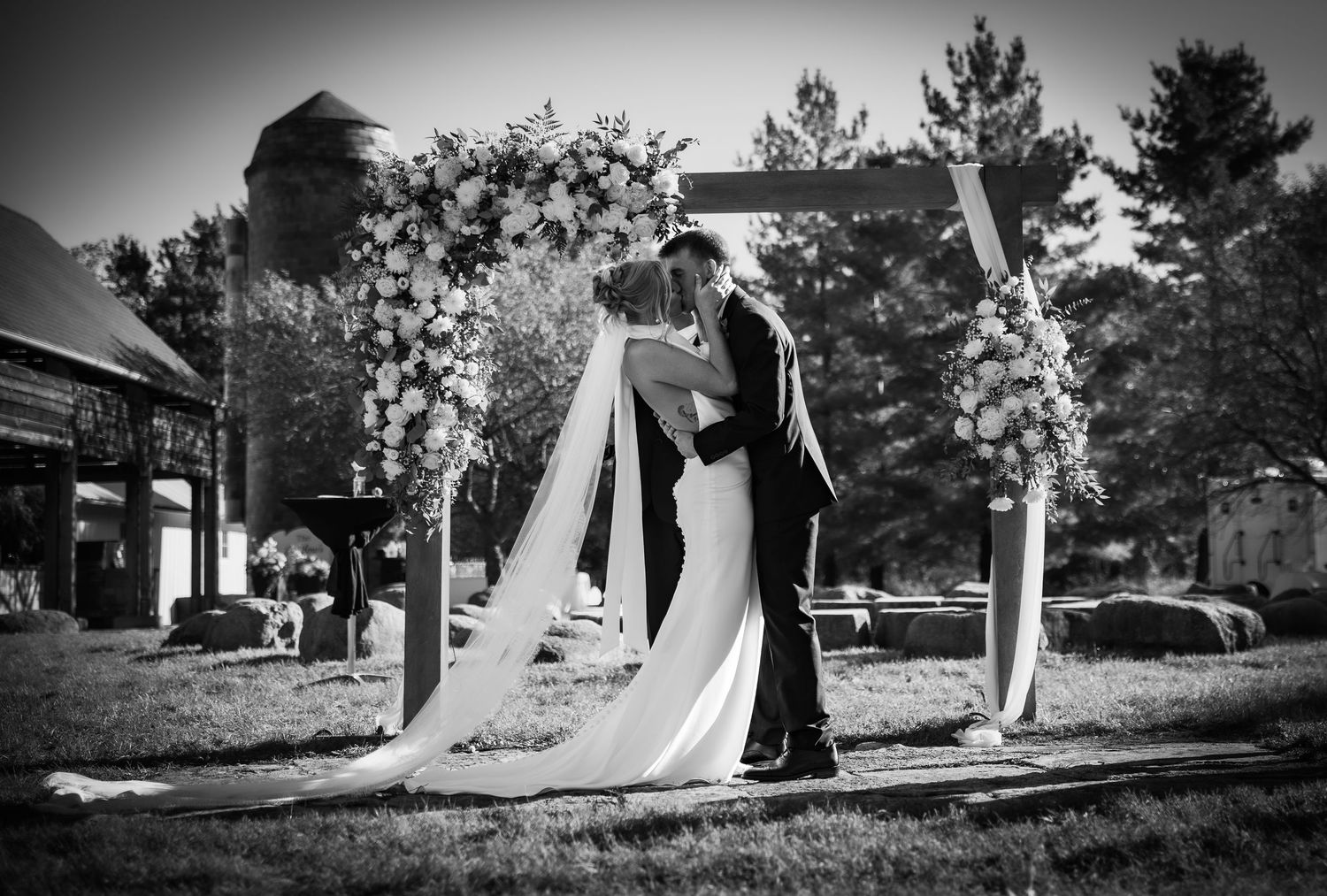 Black and white photo of a bride and groom sharing their first kiss under a floral arch at an outdoor wedding ceremony.