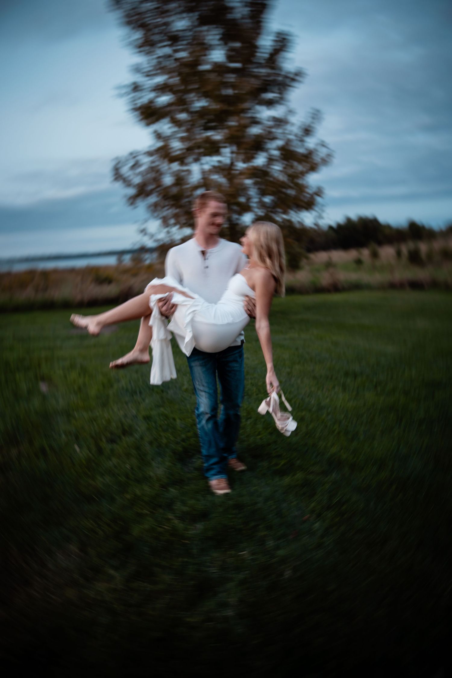 Man carrying woman in white dress across green field at dusk, romantic outdoor couple photo with soft blur effect.